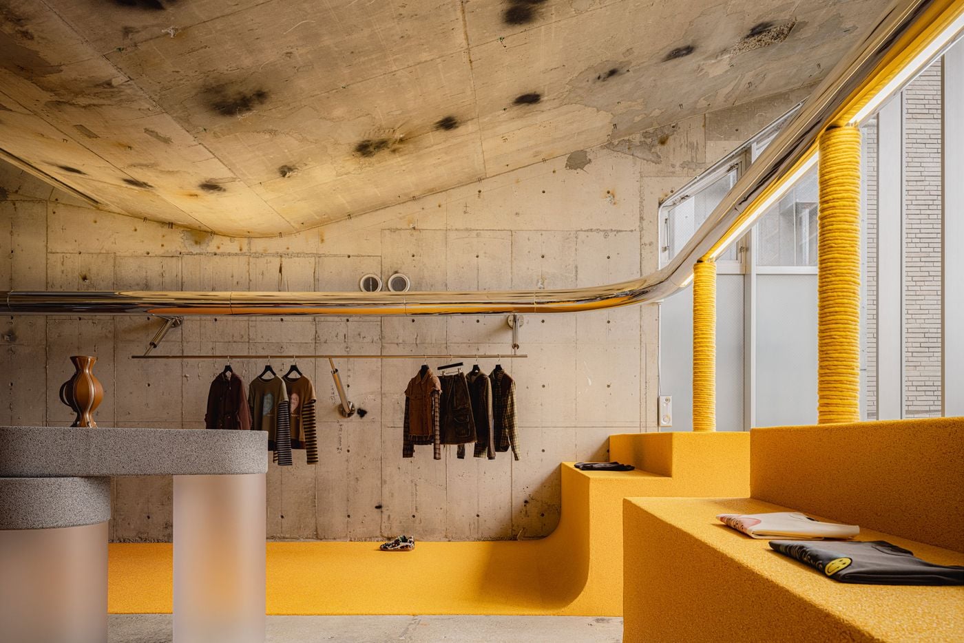 An oblique view across the upper level highlights the tension between exposed, time-marked concrete and the store’s vivid yellow interventions. Chrome pipework curves fluidly around the room and towards the windows, while rope-wrapped columns and integrated display benches bring texture and sculptural continuity to the bright, industrial interior.