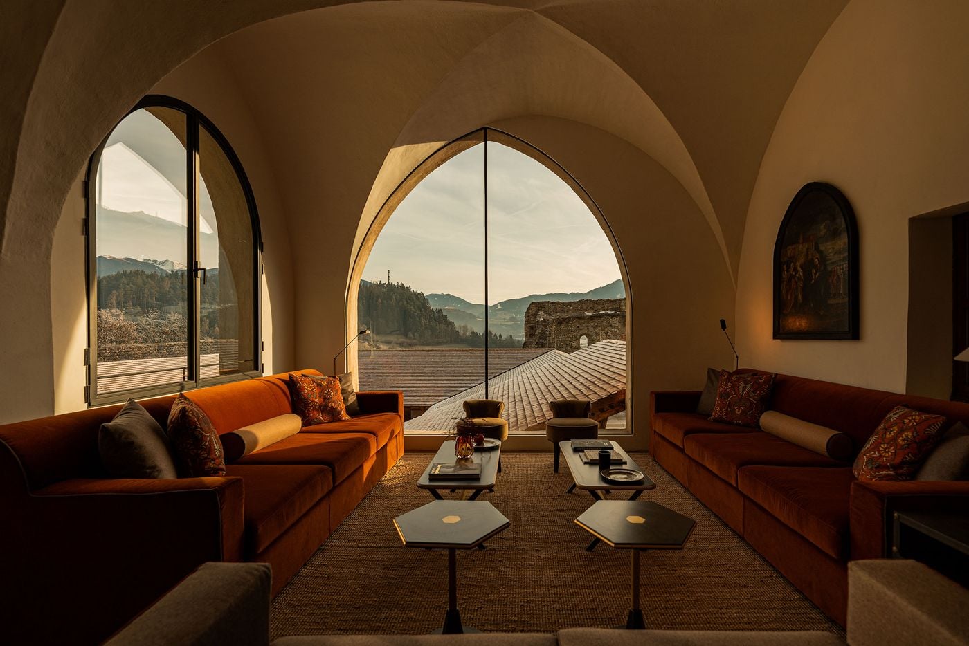 Pale groin vaults frame a frameless pointed-arch window in a Castel Badia lounge, opening onto shingled rooftops, a ruined stone wall and wooded peaks. Two facing burnt-orange velvet sofas, bolster cushions, hexagonal side tables and a jute rug lead the eye towards a pair of small slipper chairs at the glass. Pale groin vaults frame a frameless pointed-arch window in a Castel Badia lounge, opening onto shingled rooftops, a ruined stone wall and wooded peaks. Two facing burnt-orange velvet sofas, bolster cushions, hexagonal side tables and a jute rug lead the eye towards a pair of small slipper chairs at the glass.
