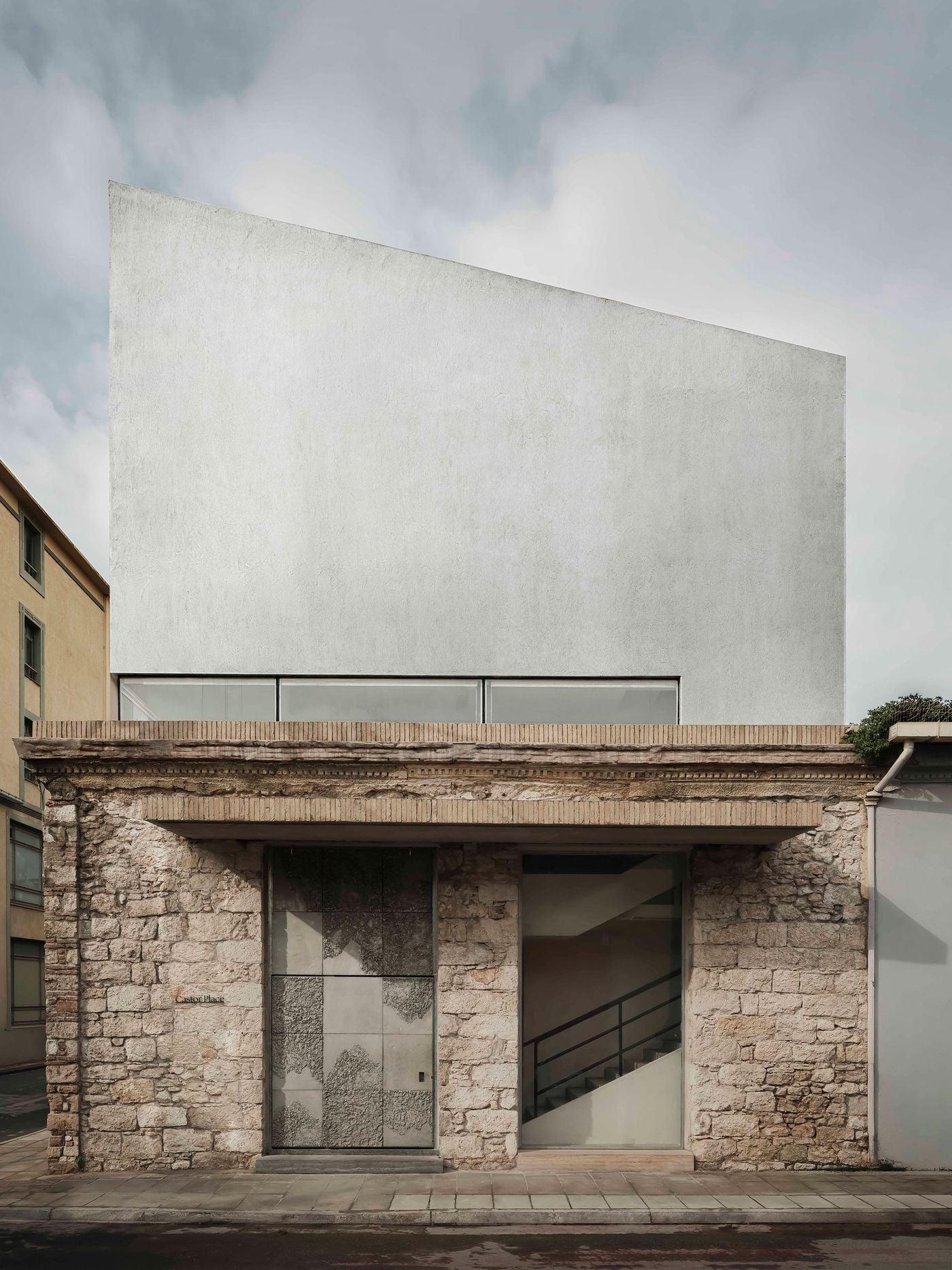 The street façade juxtaposes the low, weathered stone volume of the existing building with a tall, almost blank upper addition. A continuous strip of glazing separates old from new, while the textured entrance door and adjacent glazed opening punctuate the retained masonry at pavement level. The street façade juxtaposes the low, weathered stone volume of the existing building with a tall, almost blank upper addition. A continuous strip of glazing separates old from new, while the textured entrance door and adjacent glazed opening punctuate the retained masonry at pavement level.