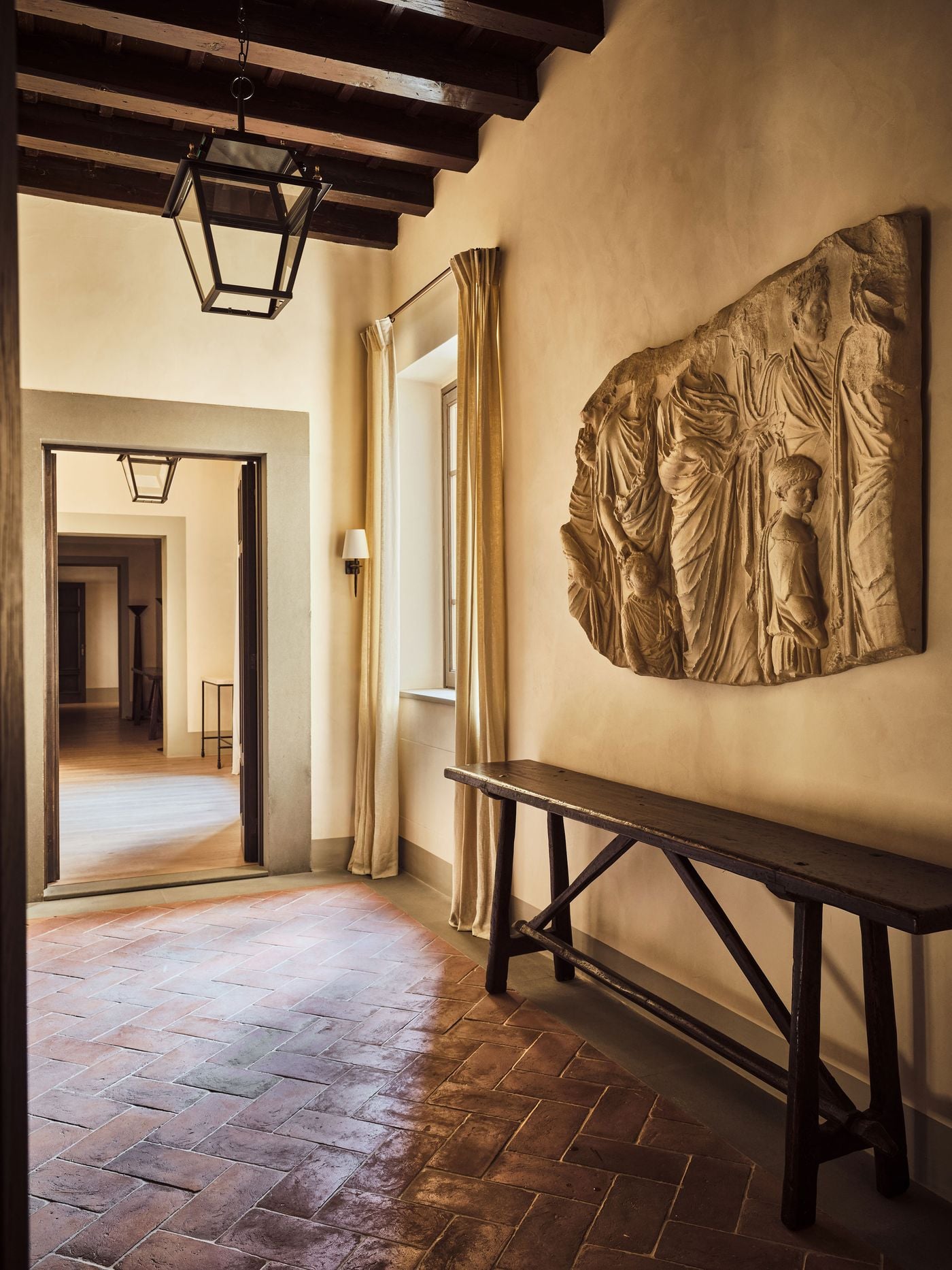 A large classical relief of robed processional figures, its edges left ragged like an excavated fragment, dominates a hallway at La Réserve Firenze. Beneath it, a rustic trestle console stands on herringbone terracotta, while dark timber beams, an iron lantern and a stone-framed doorway draw the eye through a sequence of rooms. A large classical relief of robed processional figures, its edges left ragged like an excavated fragment, dominates a hallway at La Réserve Firenze. Beneath it, a rustic trestle console stands on herringbone terracotta, while dark timber beams, an iron lantern and a stone-framed doorway draw the eye through a sequence of rooms.
