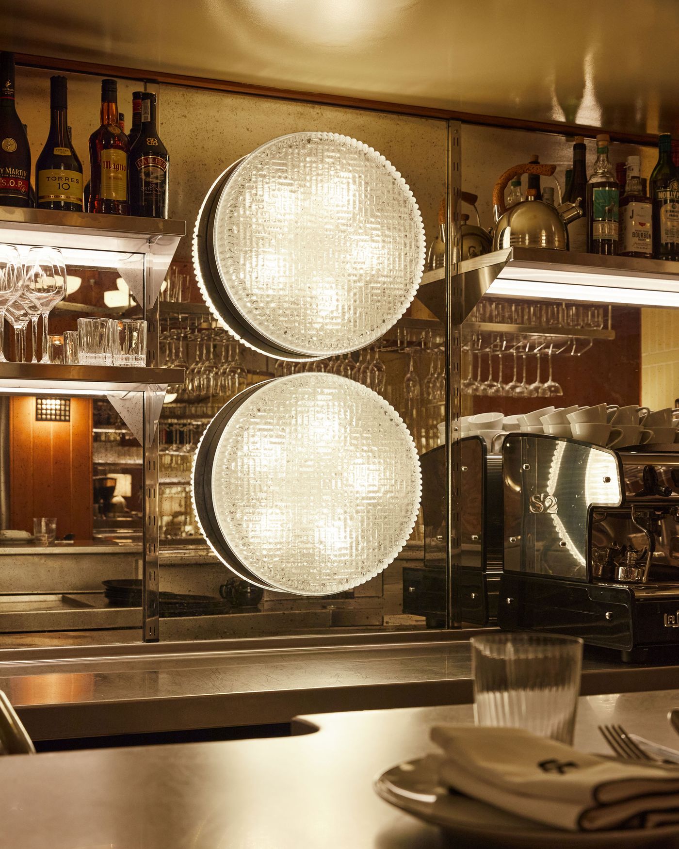 This close-up captures two prominent, circular light fixtures with a textured glass finish, hanging against a mirrored wall. The strong, direct light highlights the ribbed pattern of the glass and the reflections of bottles and glasses from the bar, creating a luminous and visually intriguing composition.