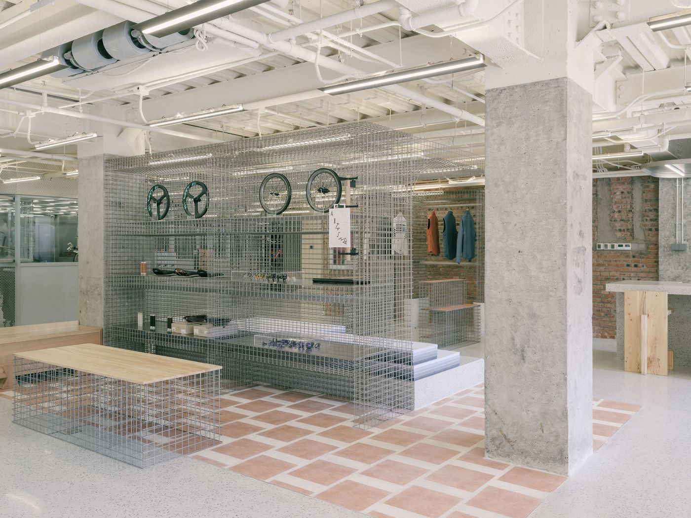 A dynamic retail display within AITASHOP features a large, light grey wire mesh structure serving as modular shelving for accessories and bicycle components. A low wooden bench element integrates into the mesh, offering seating or display space. The blend of raw concrete columns, exposed brick, and a patterned terracotta floor creates a rich, industrial-meets-minimalist aesthetic. A dynamic retail display within AITASHOP features a large, light grey wire mesh structure serving as modular shelving for accessories and bicycle components. A low wooden bench element integrates into the mesh, offering seating or display space. The blend of raw concrete columns, exposed brick, and a patterned terracotta floor creates a rich, industrial-meets-minimalist aesthetic.