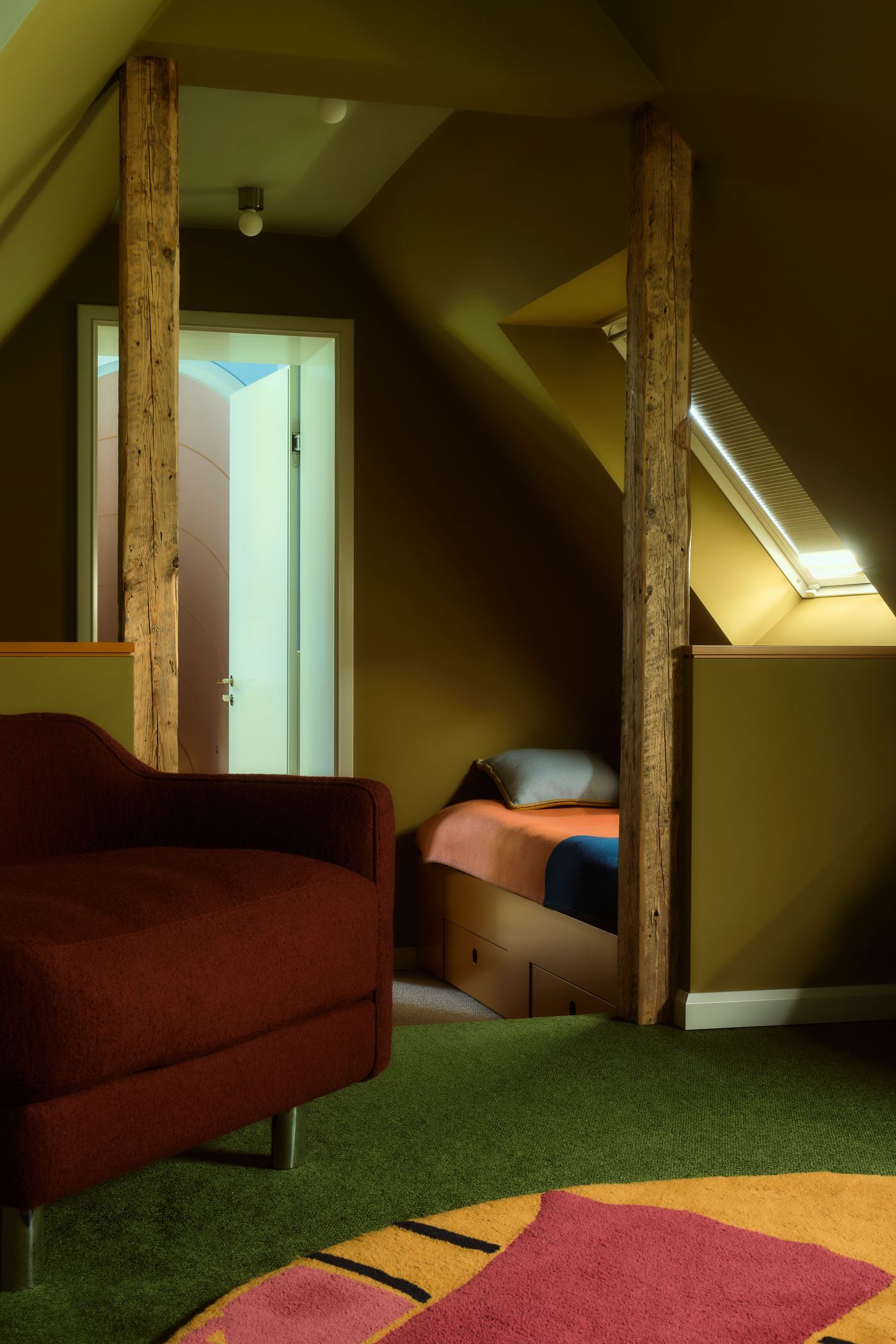 Cozy attic interior featuring a maroon chair, a colorful bed, and a vibrant rug, framed by wooden beams and a doorway. Cozy attic interior featuring a maroon chair, a colorful bed, and a vibrant rug, framed by wooden beams and a doorway.