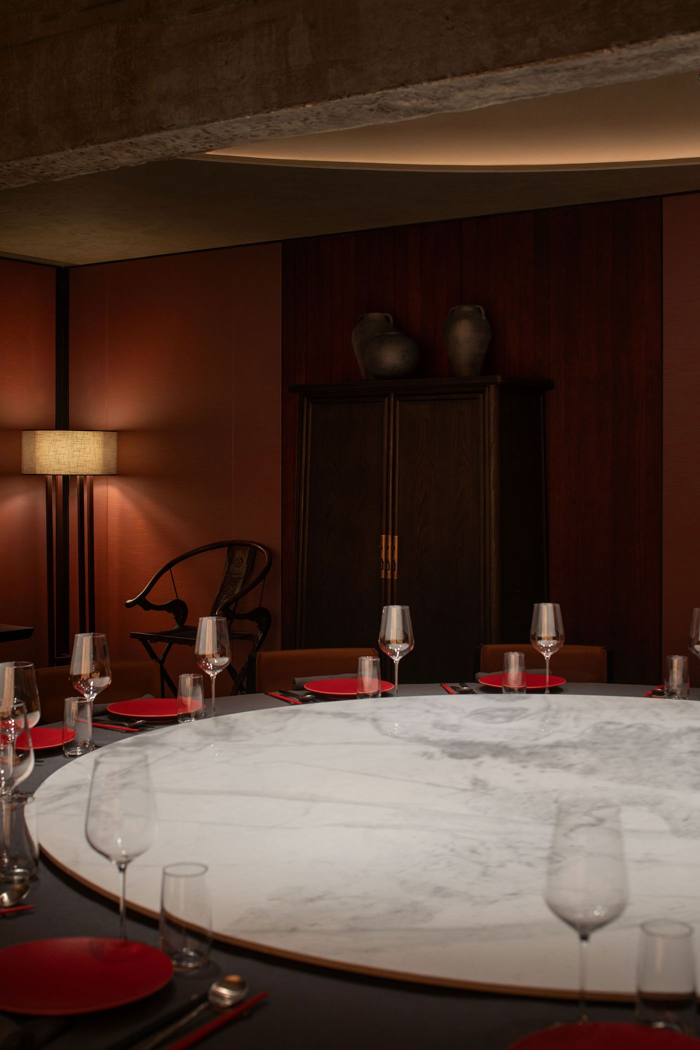 Elegant dining setup featuring a round marble table with red plates, wine glasses, and modern decor in warm, muted lighting.