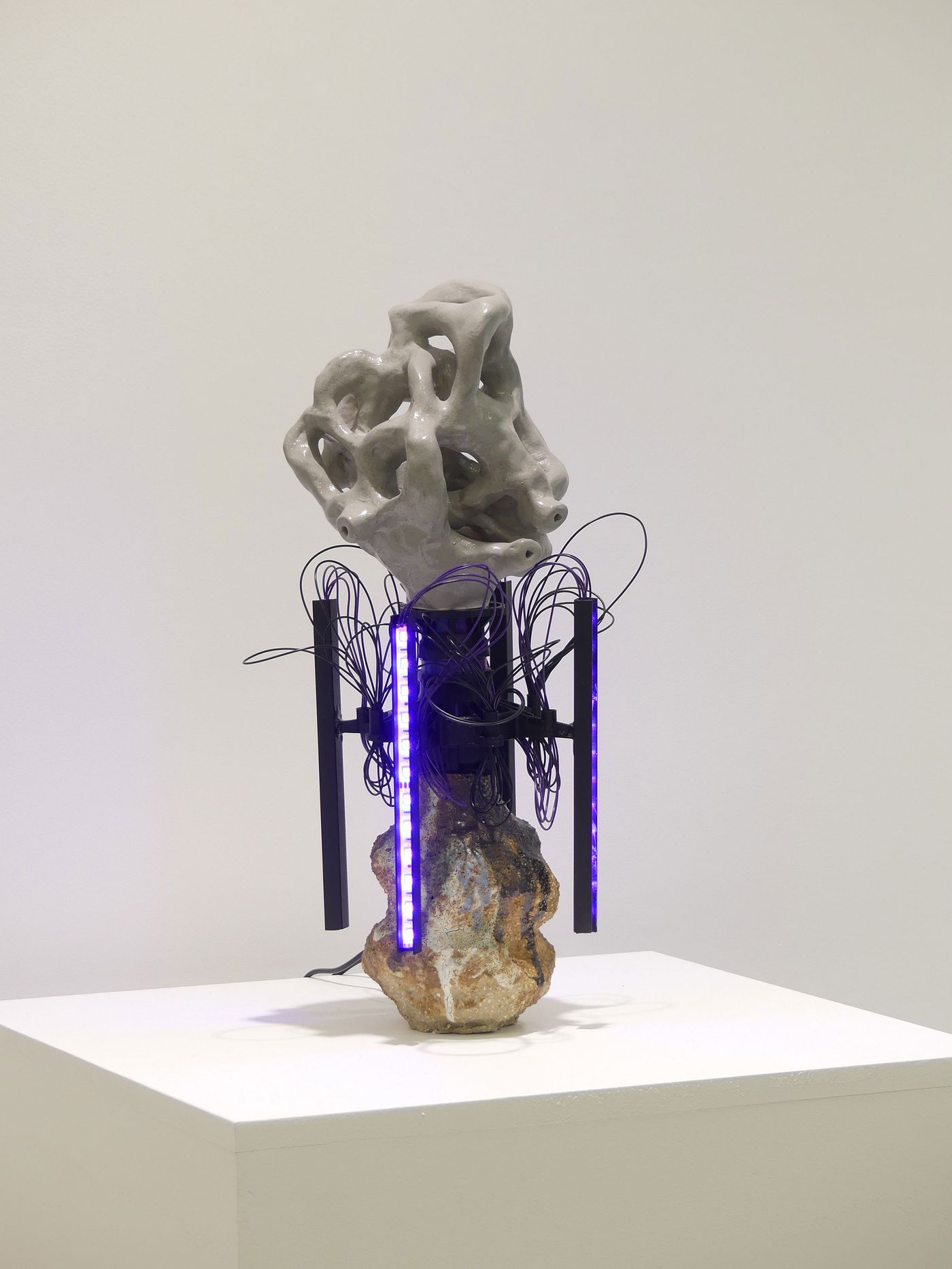A modern sculpture featuring an abstract gray form atop a textured base, surrounded by black wires and vertical purple lights.