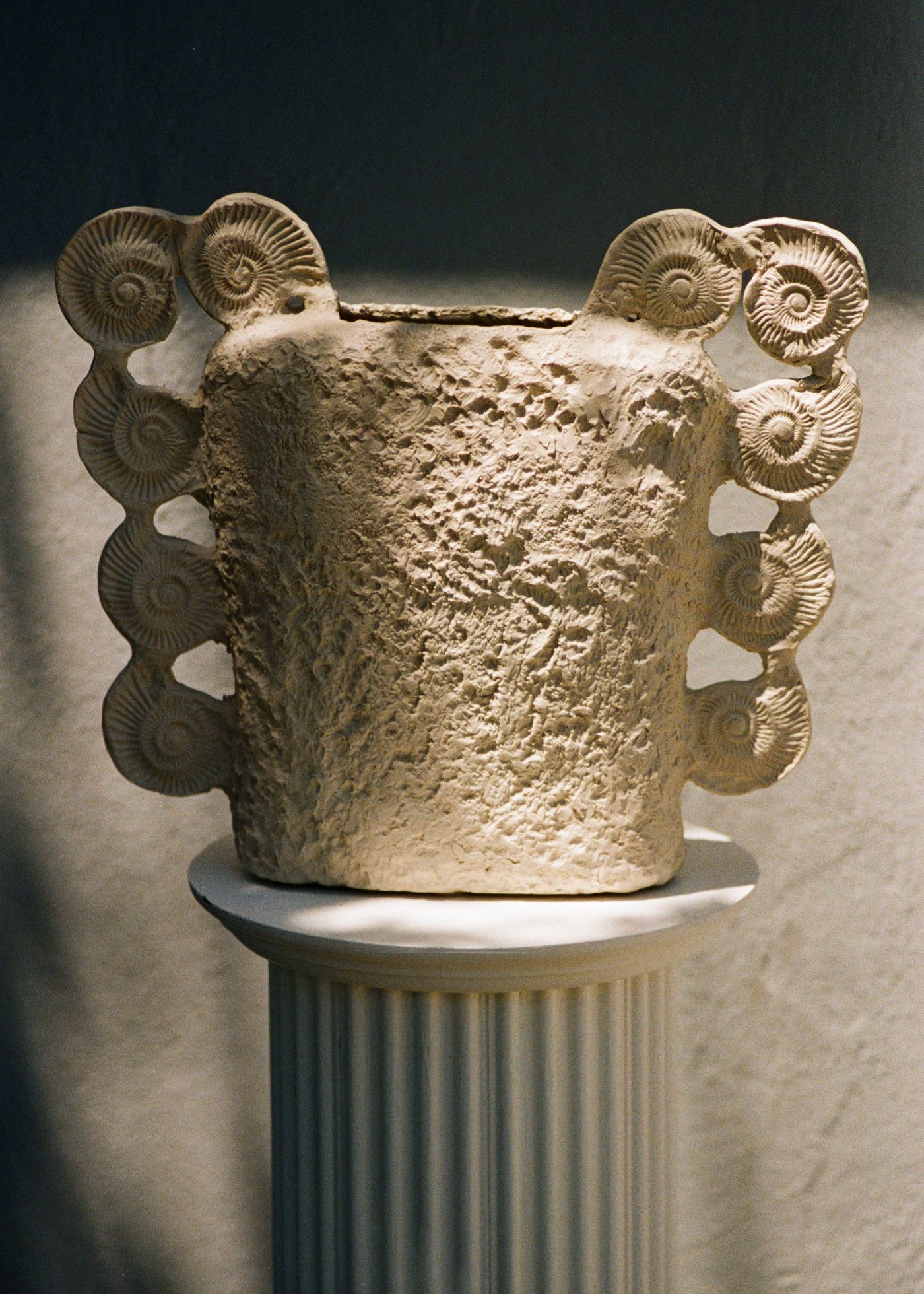 A textured, cream-colored ceramic vessel, adorned with multiple ammonite fossil imprints around its rim and sides, rests on a fluted classical column. The dramatic lighting casts strong shadows, highlighting the rough, organic texture of the pottery and the intricate detail of the fossil imprints. This piece evokes a sense of natural history and artistic craftsmanship, presented in a museum-like setting.