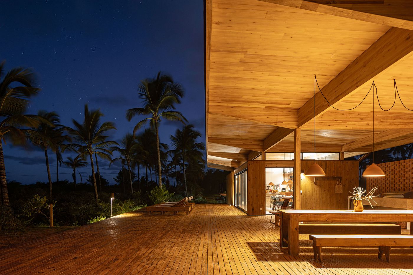An expansive wooden deck extends from a warmly lit wood-clad building under a dark, starry night sky. The soft illumination highlights the inviting outdoor dining area and lounge space, with silhouetted palm trees creating a serene, immersive tropical atmosphere. An expansive wooden deck extends from a warmly lit wood-clad building under a dark, starry night sky. The soft illumination highlights the inviting outdoor dining area and lounge space, with silhouetted palm trees creating a serene, immersive tropical atmosphere.