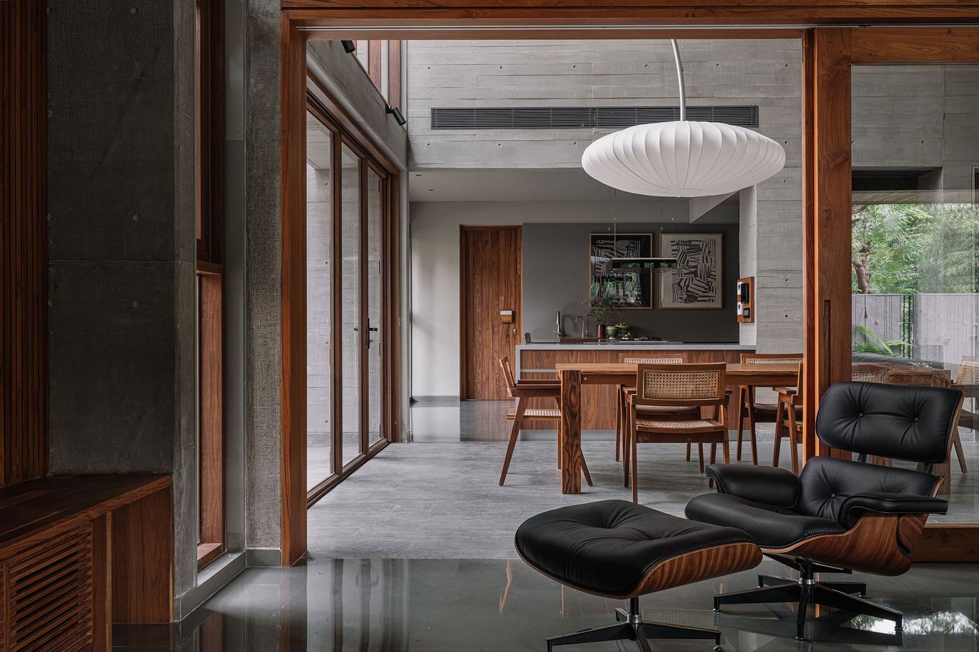 A spacious open-plan living area in the MS House, designed with raw concrete walls and a polished dark floor. A classic Eames Lounge Chair and ottoman invite relaxation. A minimalist dining table with rattan-back chairs occupies the background. A large, organic-shaped pendant light hovers, adding a soft, sculptural element to the sophisticated space.