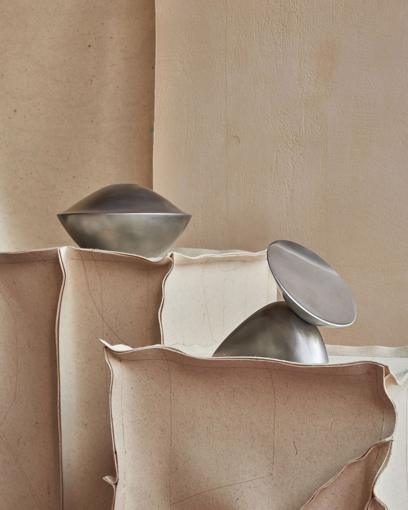 Two sleek metallic sculptures rest on textured beige paper, surrounded by soft curves and folds, blending elegance with artistry.