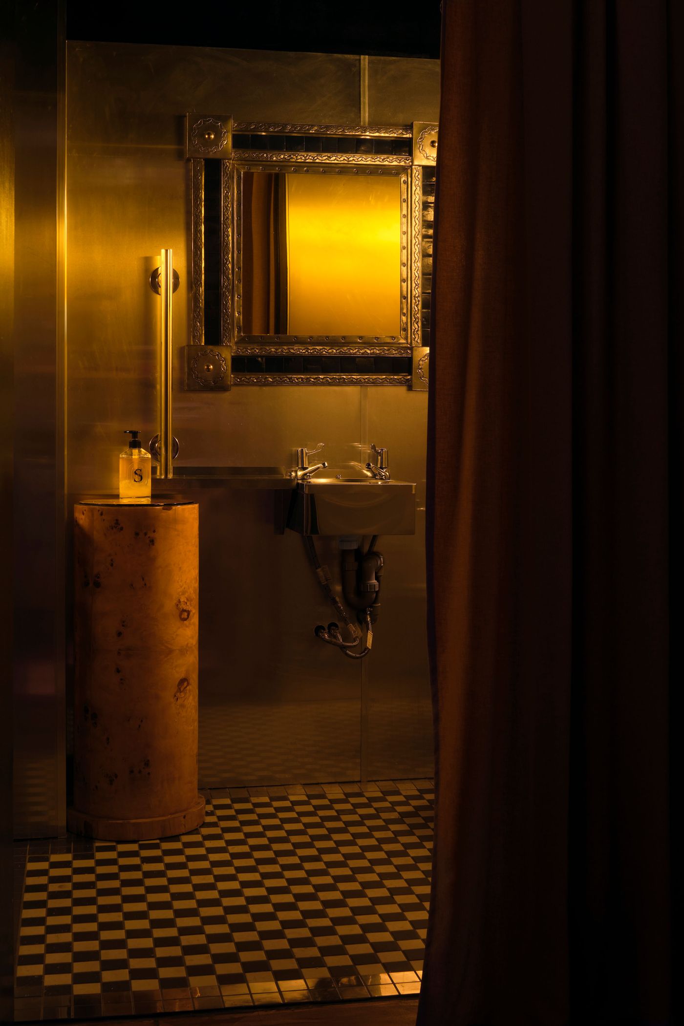 A luxurious washroom at The Roses of Elagabalus, featuring a checkerboard tiled floor and a rich, golden-toned wall. An ornate, vintage-style mirror with an intricate frame reflects the warm light, while a polished wooden pedestal with a perfume bottle stands beside a wall-mounted sink, creating an elegant and opulent Art Deco-inspired space. A luxurious washroom at The Roses of Elagabalus, featuring a checkerboard tiled floor and a rich, golden-toned wall. An ornate, vintage-style mirror with an intricate frame reflects the warm light, while a polished wooden pedestal with a perfume bottle stands beside a wall-mounted sink, creating an elegant and opulent Art Deco-inspired space.