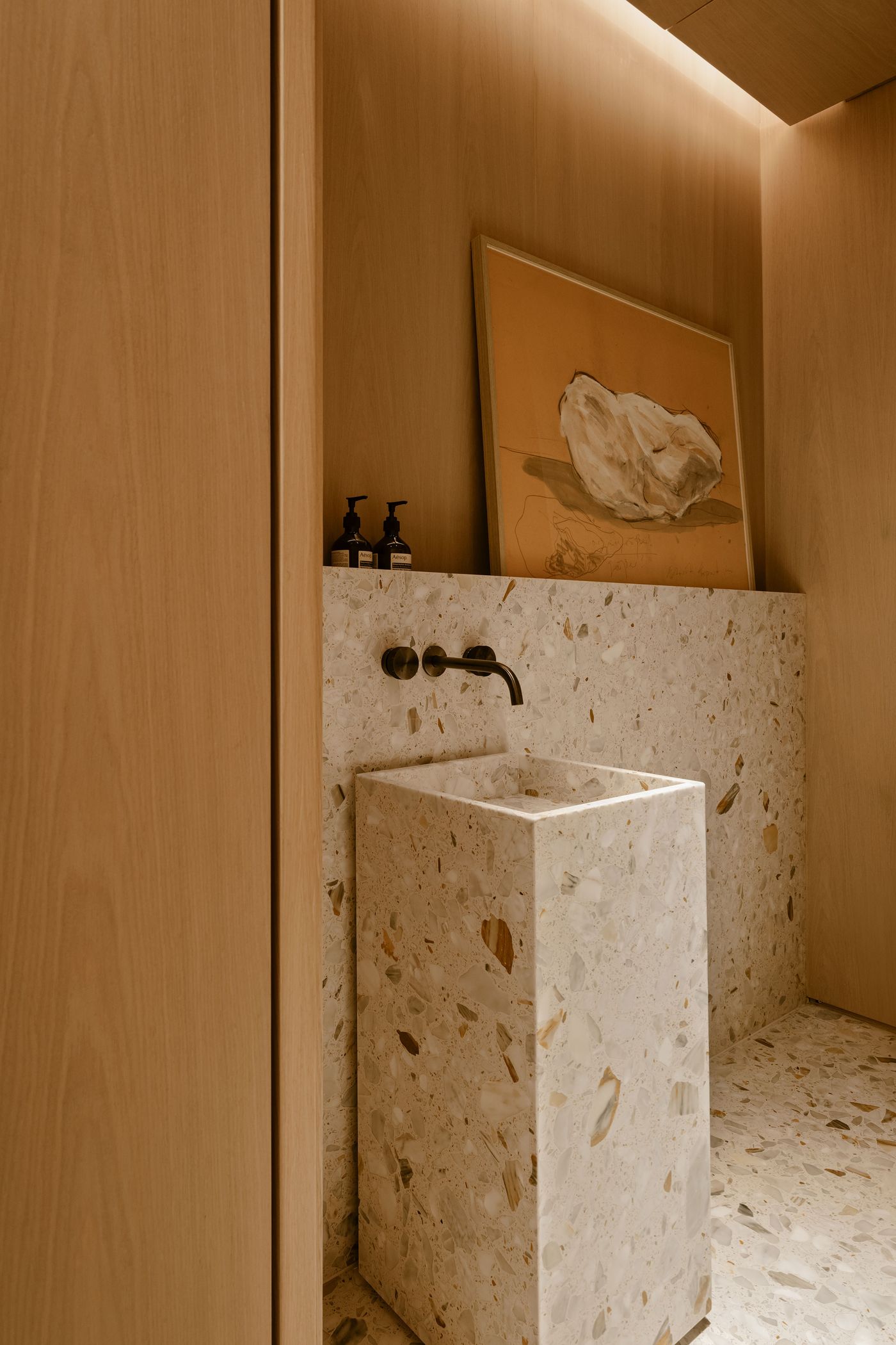 A modern bathroom features a minimalist terrazzo sink, artistic wall print, and wooden accents, creating a calm, elegant atmosphere.