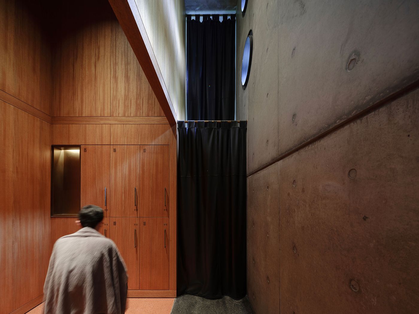 A person in a gray blanket walks past wooden storage cabinets, a black curtain, and a concrete wall with round windows above. A person in a gray blanket walks past wooden storage cabinets, a black curtain, and a concrete wall with round windows above.