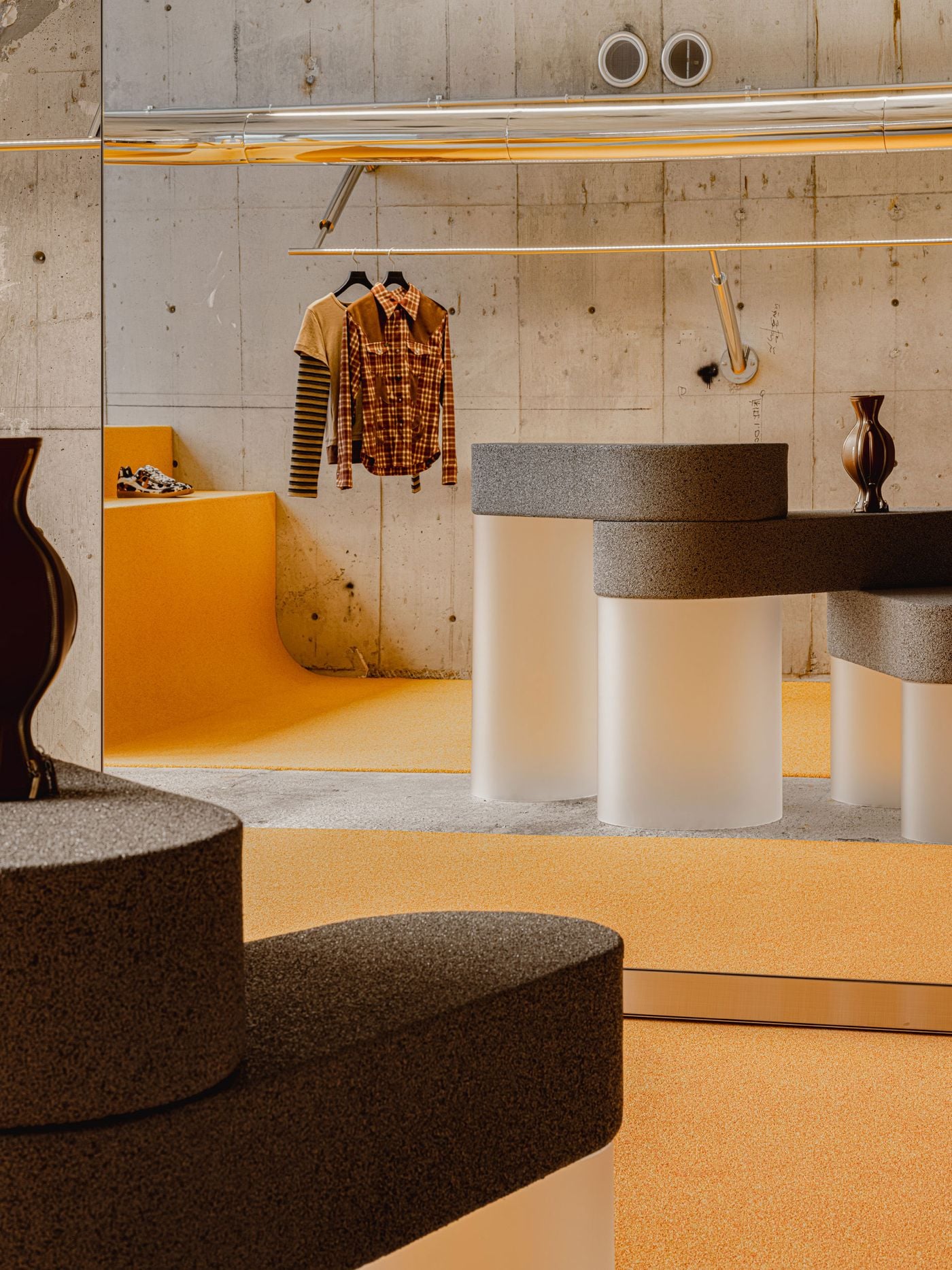 Layered display elements animate the upper floor of ANDERSSON BELL Tokyo, where yellow microcement, raw concrete and chrome rails frame clothing against a highly controlled backdrop. A sculptural grey counter on pale cylindrical supports introduces rounded forms, while mirrored surfaces multiply the store’s contrasting textures and colours.