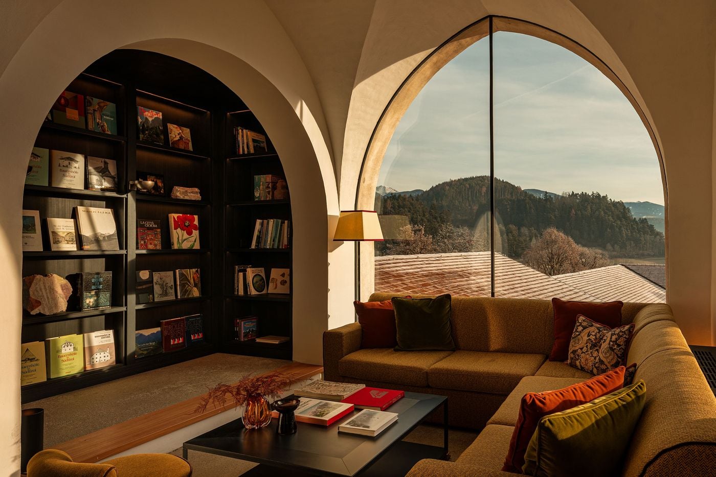A deep arched niche at Castel Badia holds dark-stained shelving lined with books on South Tyrolean farmhouses, castles and Alpine art, alongside rough mineral specimens. Opposite, an ochre corner sofa piled with olive and russet cushions faces a towering pointed-arch window onto forested hills and snow-capped summits. A deep arched niche at Castel Badia holds dark-stained shelving lined with books on South Tyrolean farmhouses, castles and Alpine art, alongside rough mineral specimens. Opposite, an ochre corner sofa piled with olive and russet cushions faces a towering pointed-arch window onto forested hills and snow-capped summits.