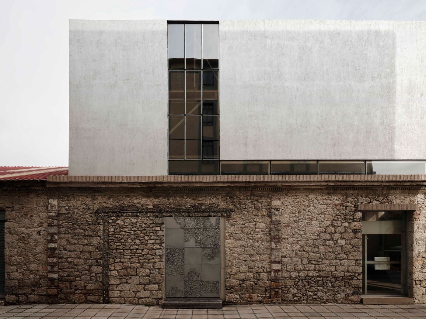 A wider elevation of Castor Place reveals the contrast between the building’s irregular historic masonry and the restrained geometry of MPNYC’s upper addition. Horizontal ribbon windows and a tall vertical glazed opening cut through the pale textured volume, while the original stone façade remains prominently legible below. A wider elevation of Castor Place reveals the contrast between the building’s irregular historic masonry and the restrained geometry of MPNYC’s upper addition. Horizontal ribbon windows and a tall vertical glazed opening cut through the pale textured volume, while the original stone façade remains prominently legible below.