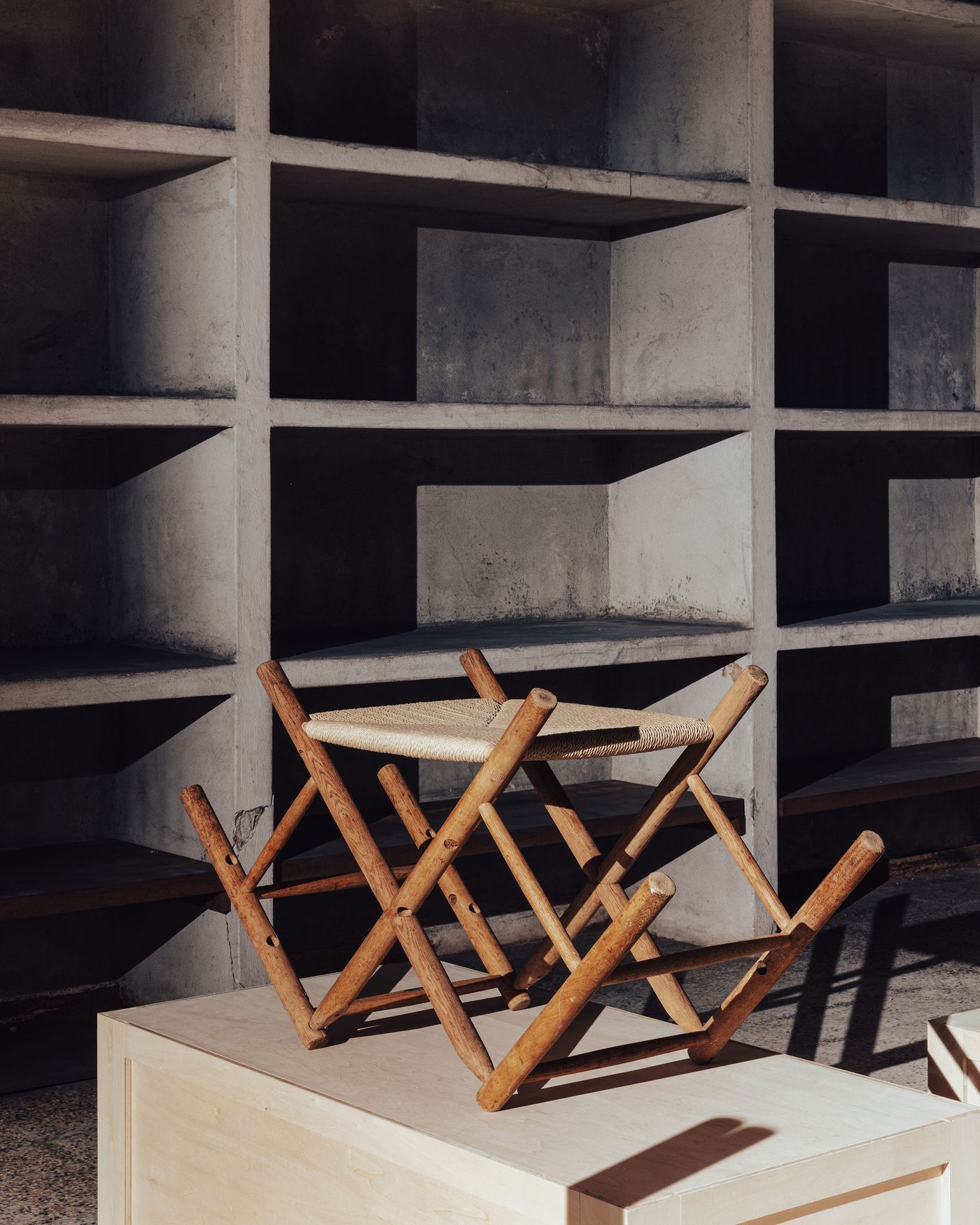 A wooden folding stool with a woven seat is displayed on a wooden platform in a minimalist space with empty concrete shelves. A wooden folding stool with a woven seat is displayed on a wooden platform in a minimalist space with empty concrete shelves.