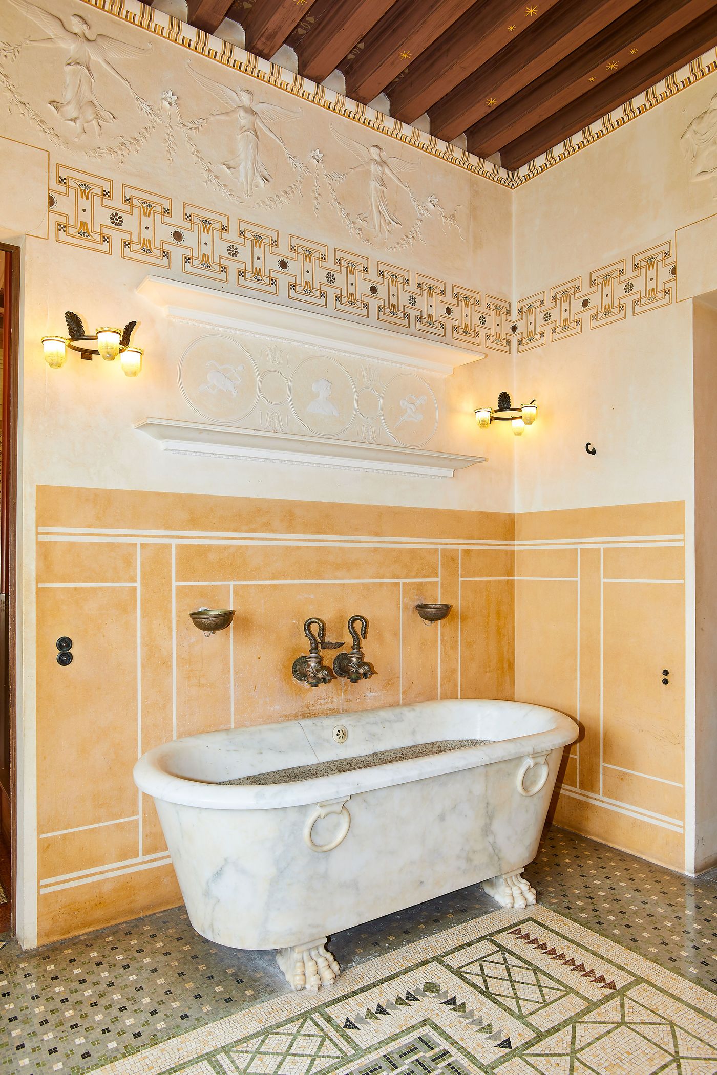 A wide shot of a Grecian-styled bathroom. A freestanding white marble bathtub with carved animalistic feet sits on an intricate mosaic tile floor. The walls are a warm, golden hue, decorated with white plaster reliefs and geometric friezes. Bronze sconces provide warm, ambient light, highlighting the fusion of ancient Greek and Roman aesthetics with contemporary design. A wide shot of a Grecian-styled bathroom. A freestanding white marble bathtub with carved animalistic feet sits on an intricate mosaic tile floor. The walls are a warm, golden hue, decorated with white plaster reliefs and geometric friezes. Bronze sconces provide warm, ambient light, highlighting the fusion of ancient Greek and Roman aesthetics with contemporary design.