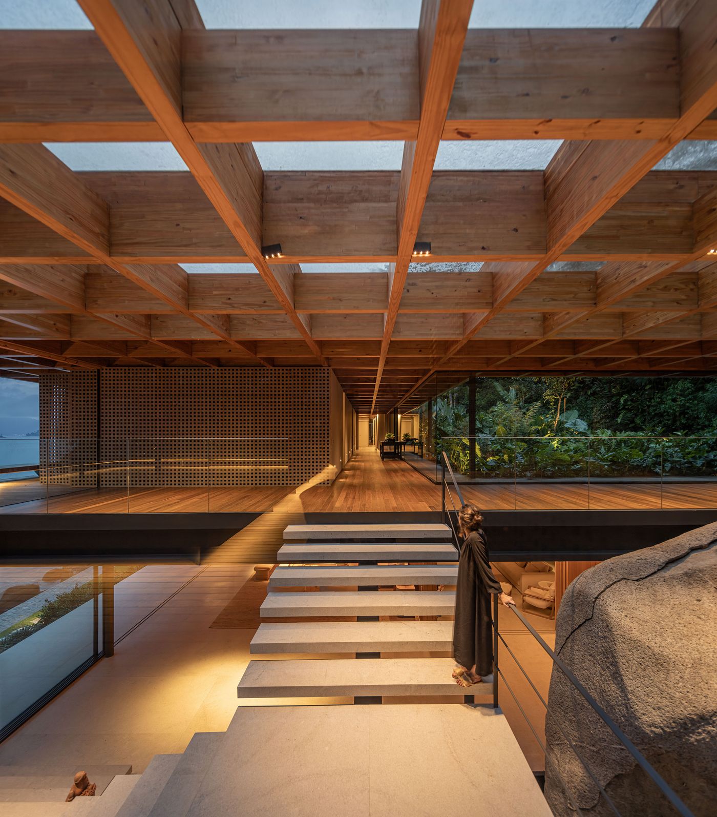A breathtaking interior view showcases a grand wooden grid ceiling with skylights, illuminating a double-height space. A floating concrete staircase gracefully ascends beside an integrated natural boulder, while a large glass wall reveals expansive views of the distant ocean and lush rainforest, embodying a sophisticated biophilic design.