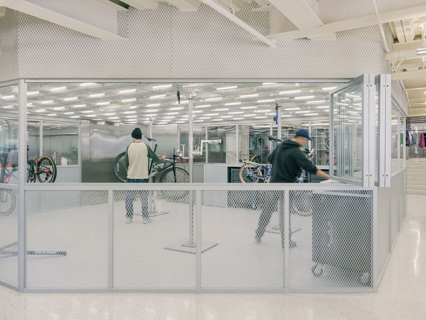 The transparent bicycle workshop allows full visibility into the mechanics' work, enclosed by clear glass and a lower band of perforated metal. Two technicians are engaged in bike assembly and maintenance, underscoring the brand's dedication to service and craftsmanship within a highly functional and minimalist industrial setting, under bright, organized lighting. The transparent bicycle workshop allows full visibility into the mechanics' work, enclosed by clear glass and a lower band of perforated metal. Two technicians are engaged in bike assembly and maintenance, underscoring the brand's dedication to service and craftsmanship within a highly functional and minimalist industrial setting, under bright, organized lighting.
