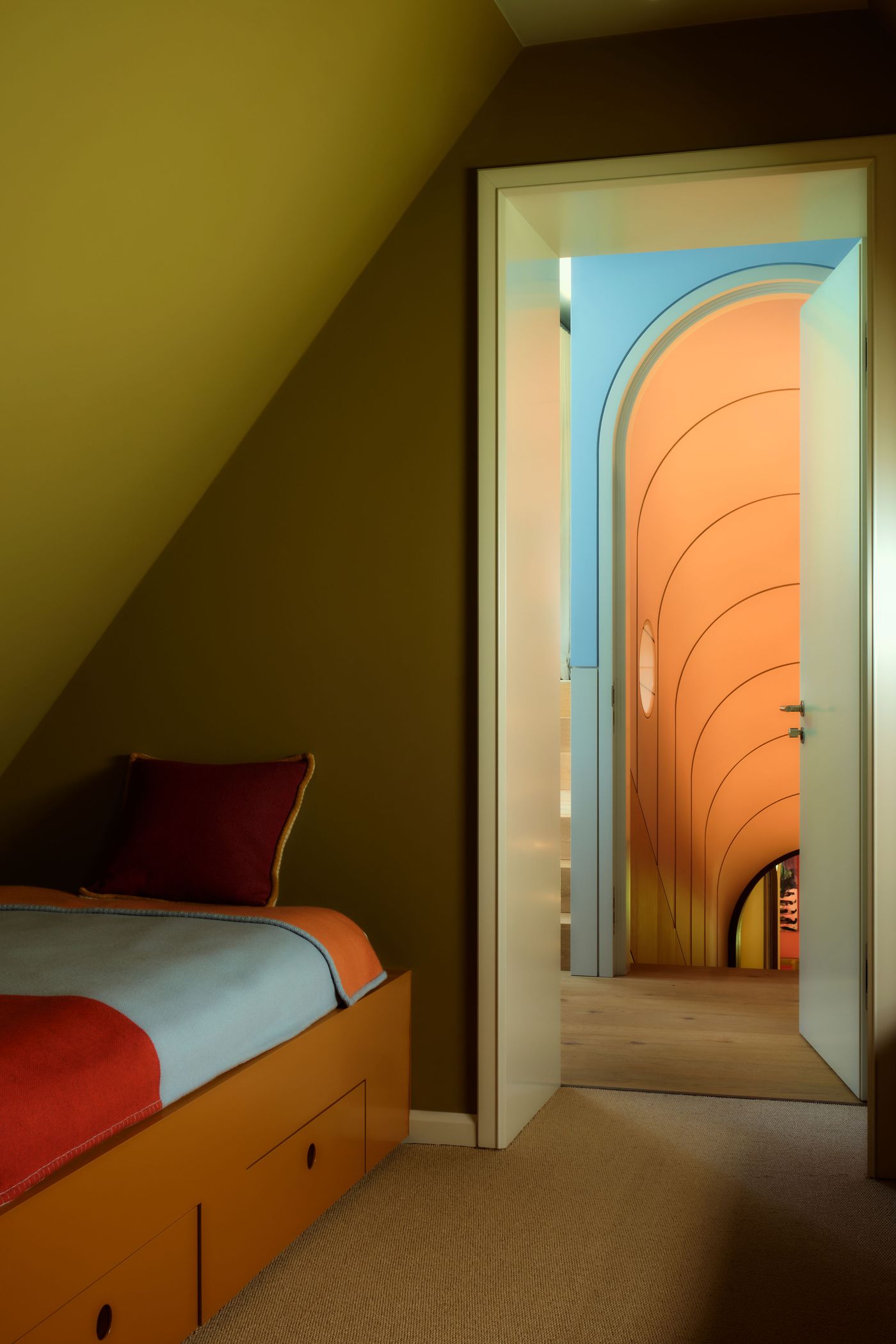 View through a doorway showcasing a cozy bedroom with an orange bedspread, leading to a colorful archway and vibrant hallway.