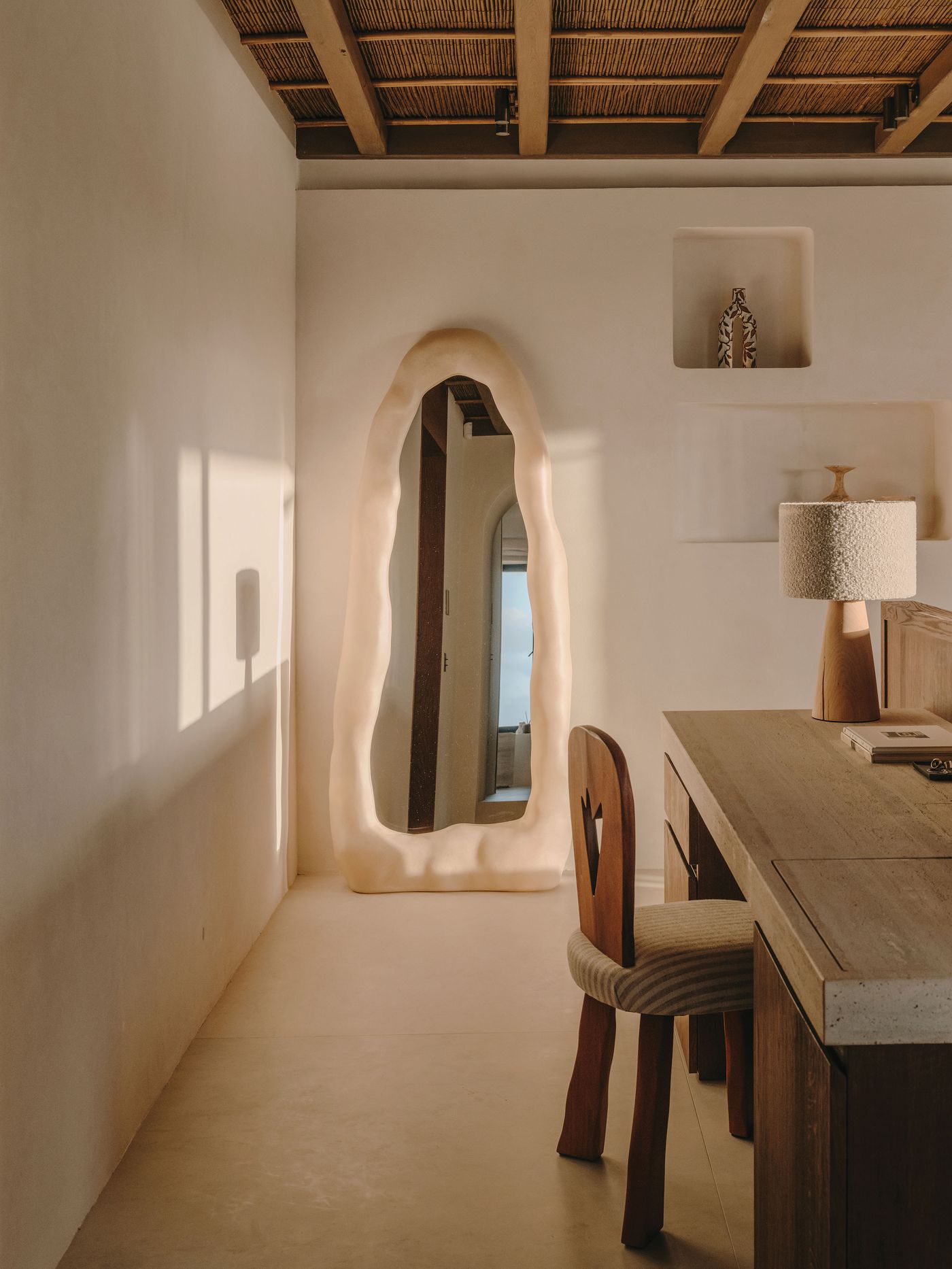 An organic-shaped, sculptural mirror leans against a cream wall in a minimalist room featuring a wooden desk and a textured lamp. An organic-shaped, sculptural mirror leans against a cream wall in a minimalist room featuring a wooden desk and a textured lamp.
