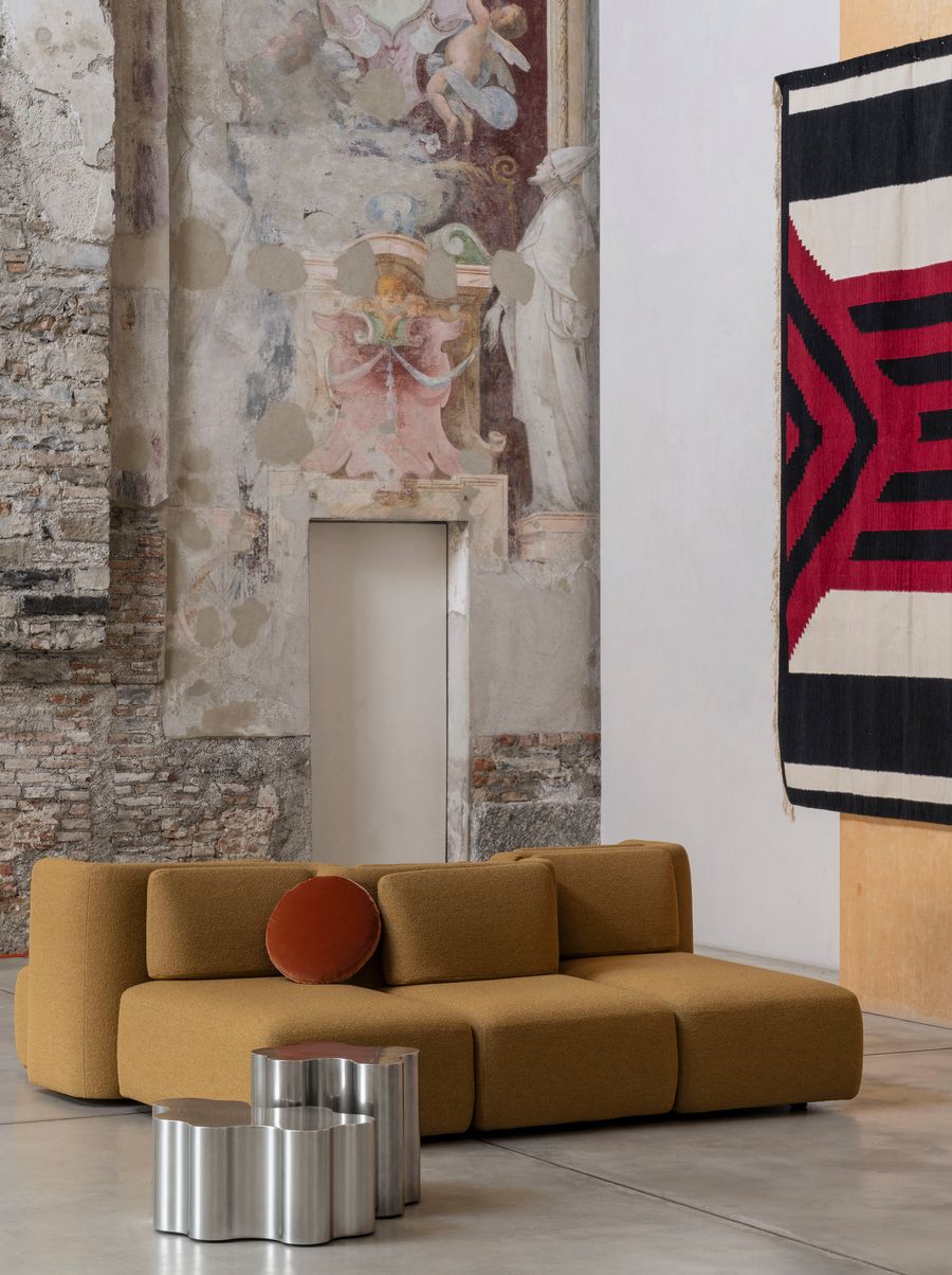 A mustard-yellow modular sofa with a textured boucle fabric is the centerpiece of this room, complemented by a round rust-colored accent pillow. Two sculptural silver side tables with a flowing, wave-like design sit beside it. The background reveals a historic wall with faded frescoes and a striking black, white, and red woven textile hanging on the wall, creating an eclectic fusion of contemporary comfort and historical grandeur.