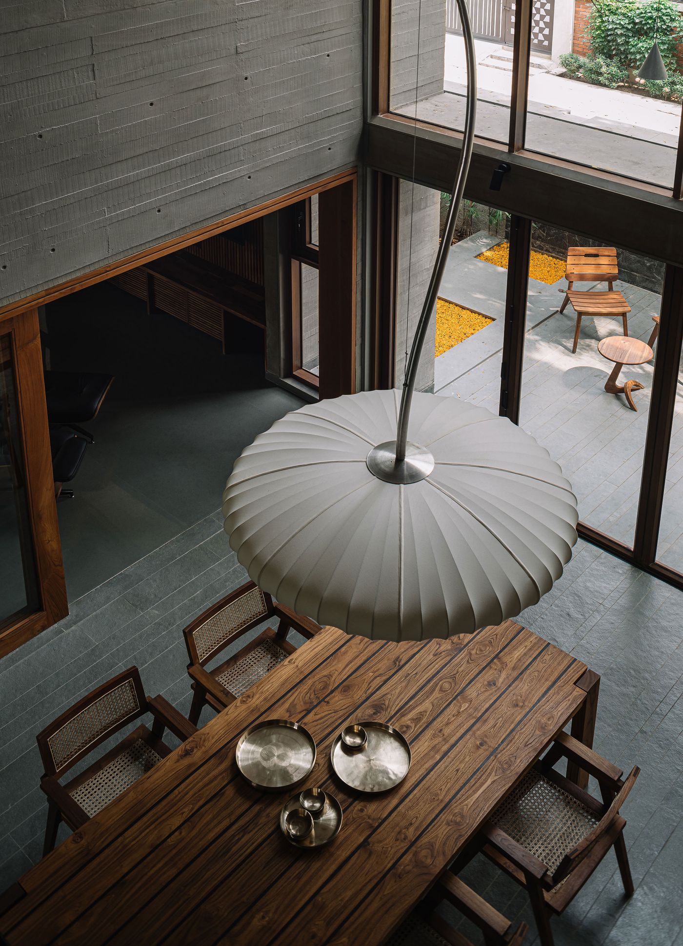 An overhead view of the dining area in the MS House, highlighting a large, textured paper lantern hanging above a substantial wooden dining table with rattan-back chairs. The room features a dark tiled floor and raw concrete walls, while expansive glass doors offer a glimpse into an outdoor space with yellow stone chippings, creating a blend of earthy and minimalist design.