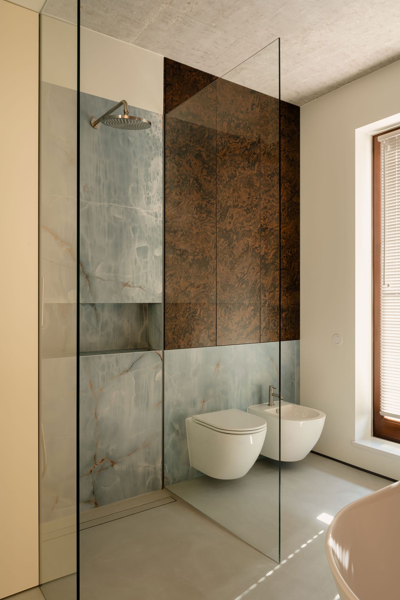 A glass-partitioned bathroom pairs soft grey terrazzo flooring with sky-blue onyx walls and a walnut veneer accent, framing a minimalist walk-in shower, wall-mounted toilet, and bidet in a serene composition.