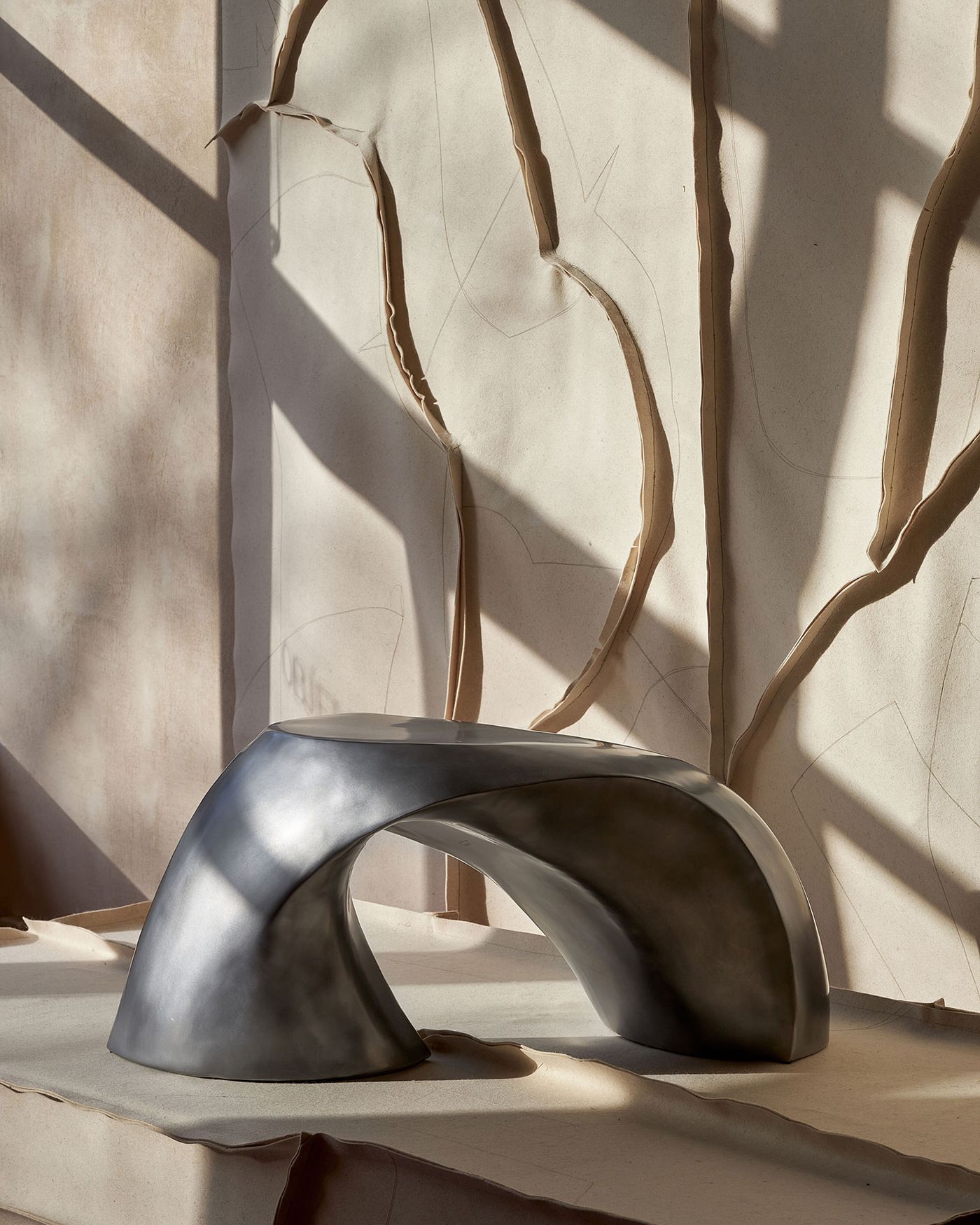 A sculptural, metallic table with a fluid design sits on textured beige surface, illuminated by soft shadows from a nearby window.
