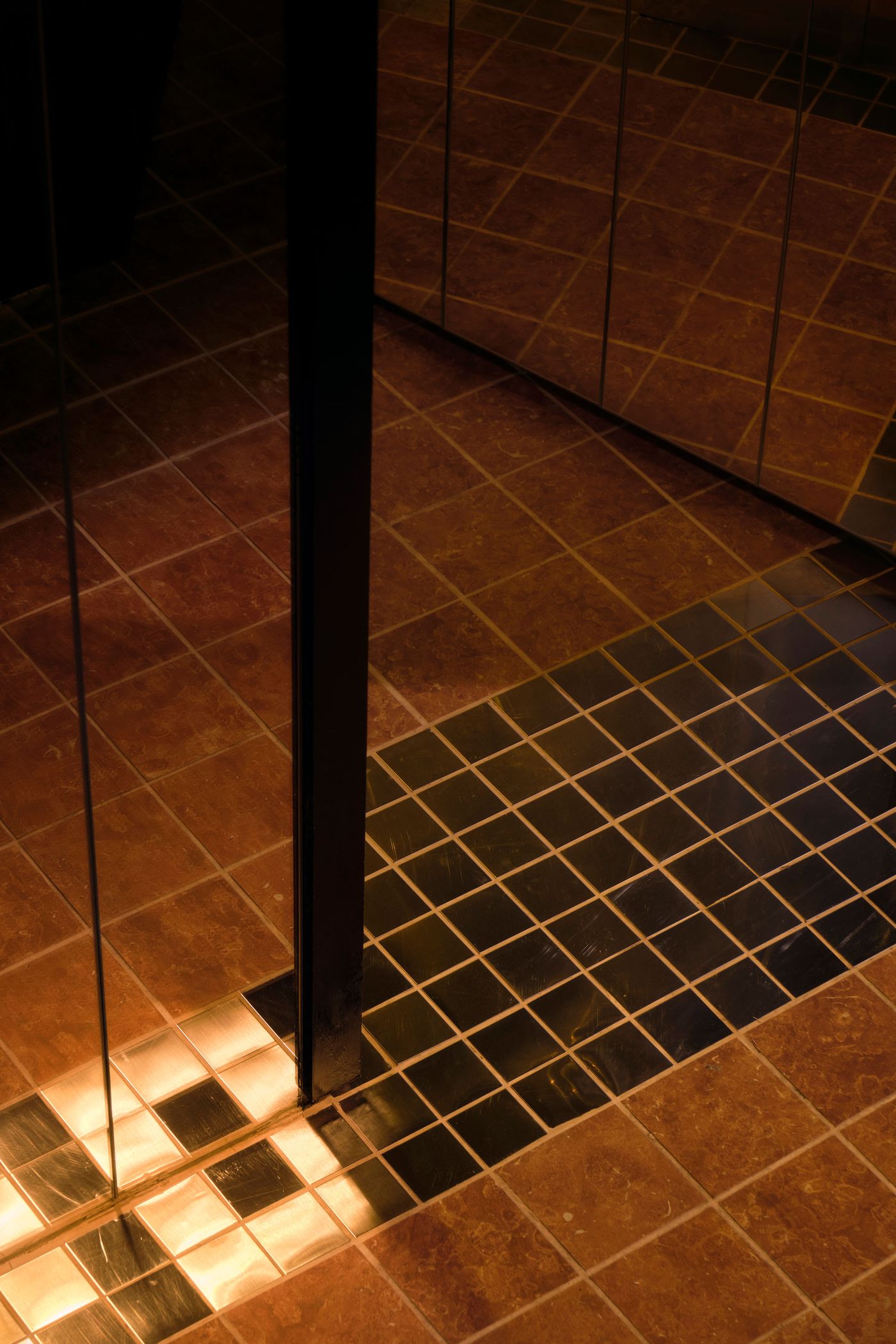 A captivating view of the floor in The Roses of Elagabalus, featuring a geometric pattern of terracotta and black tiles that reflect the ambient light. A dark, reflective vertical surface, possibly a mirrored column or wall, intersects the frame, creating a sense of depth and mystery, and enhancing the opulent, vintage-inspired design.