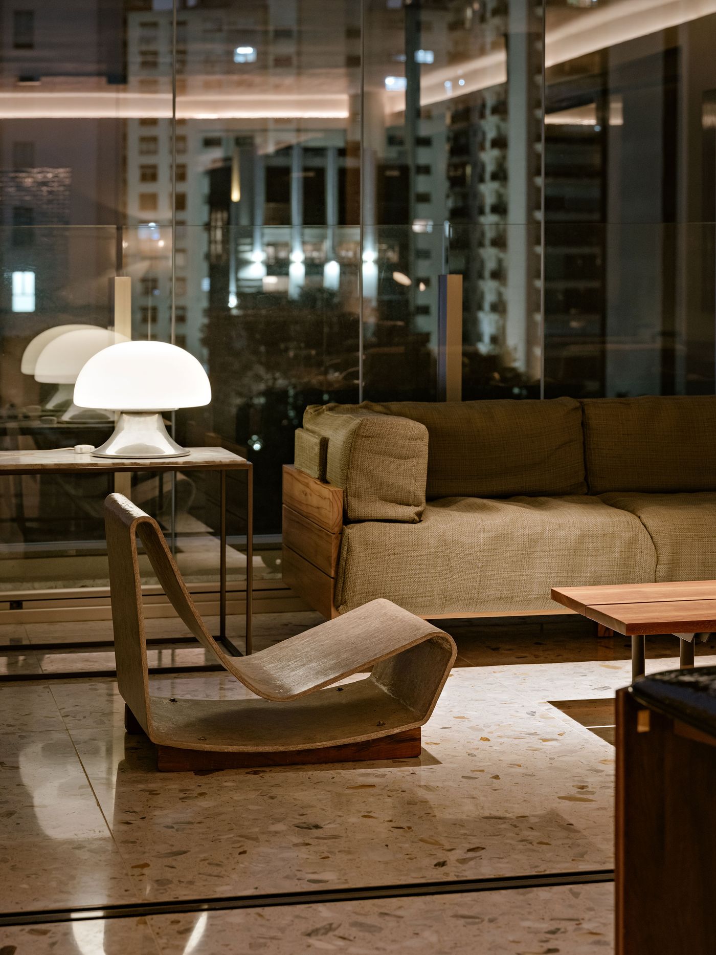 A modern living space featuring a cozy sofa, a unique curved chair, and a stylish lamp, all set against a city night backdrop.