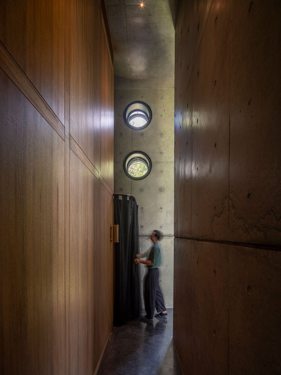 A person stands in a narrow corridor between wooden and concrete walls, with round windows letting in natural light above. A person stands in a narrow corridor between wooden and concrete walls, with round windows letting in natural light above.