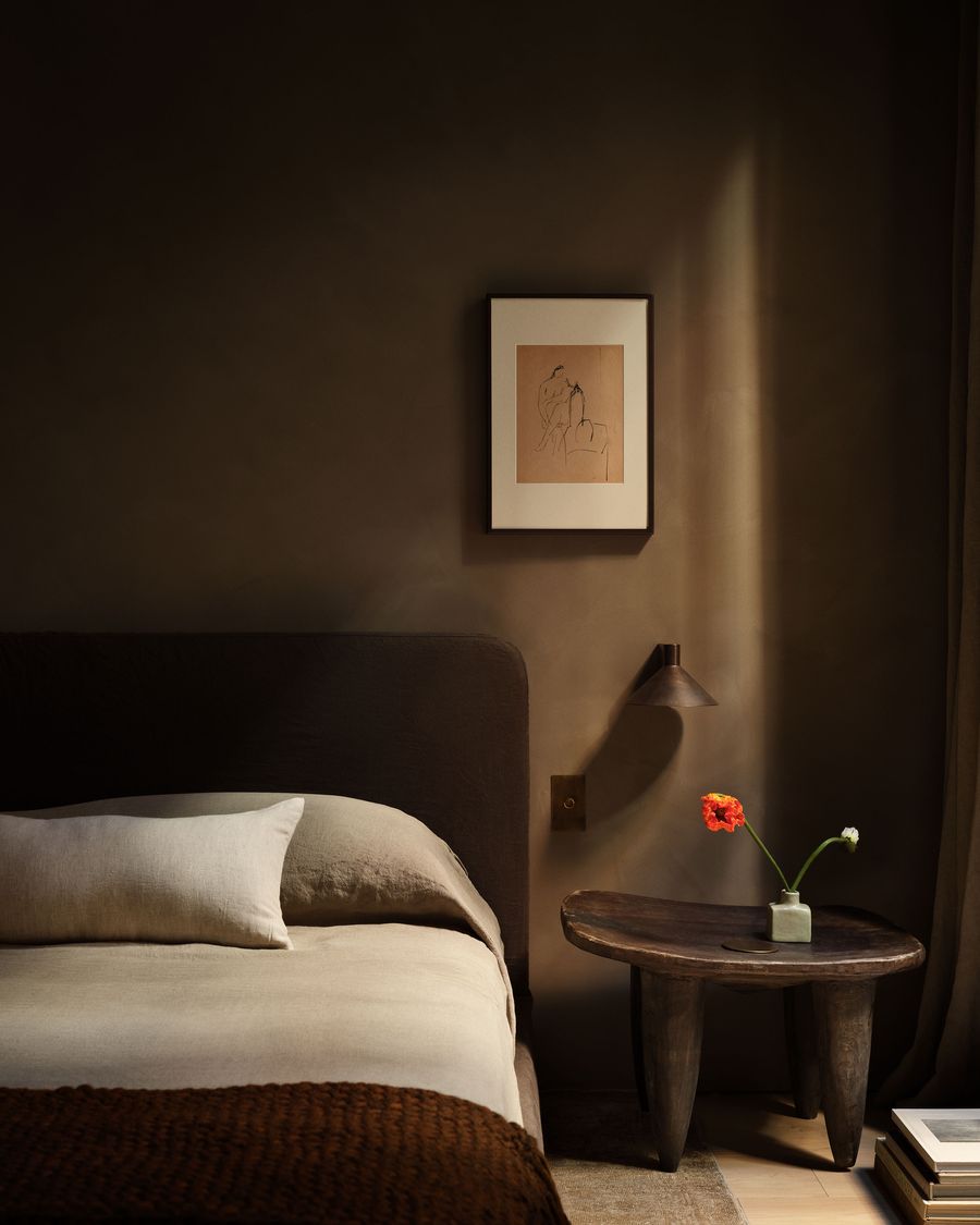 A cozy bedroom featuring a dark bed, wooden side table with a flower, and a minimalist drawing on the wall illuminated by soft light. A cozy bedroom featuring a dark bed, wooden side table with a flower, and a minimalist drawing on the wall illuminated by soft light.