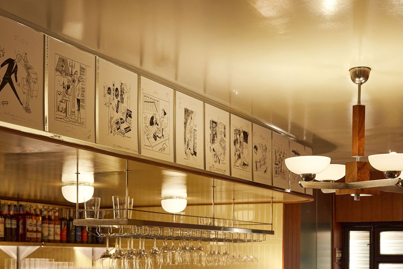 This interior view focuses on the upper bar area, where a line of whimsical, black-and-white drawings adds an element of nostalgic Art Deco flair. Below, a sleek, modern bar light fixture holds a rack of elegant glassware. The glossy ceiling reflects the light, enhancing the warm and sophisticated atmosphere of the space. This interior view focuses on the upper bar area, where a line of whimsical, black-and-white drawings adds an element of nostalgic Art Deco flair. Below, a sleek, modern bar light fixture holds a rack of elegant glassware. The glossy ceiling reflects the light, enhancing the warm and sophisticated atmosphere of the space.