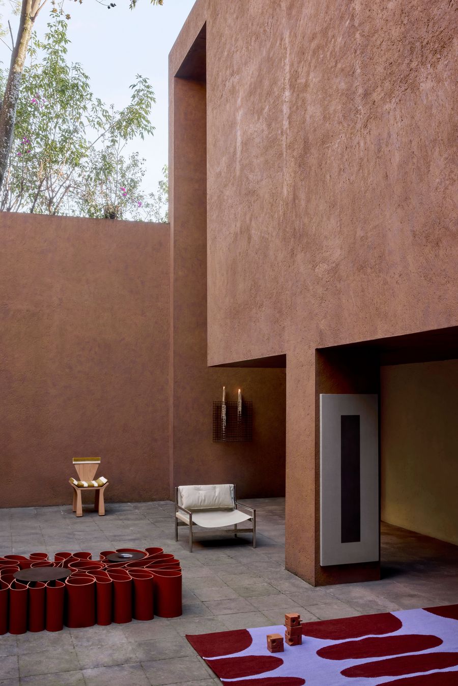 Stylish outdoor space with earthy walls, minimalist furniture, a vibrant rug, and decorative elements, surrounded by trees and soft lighting.