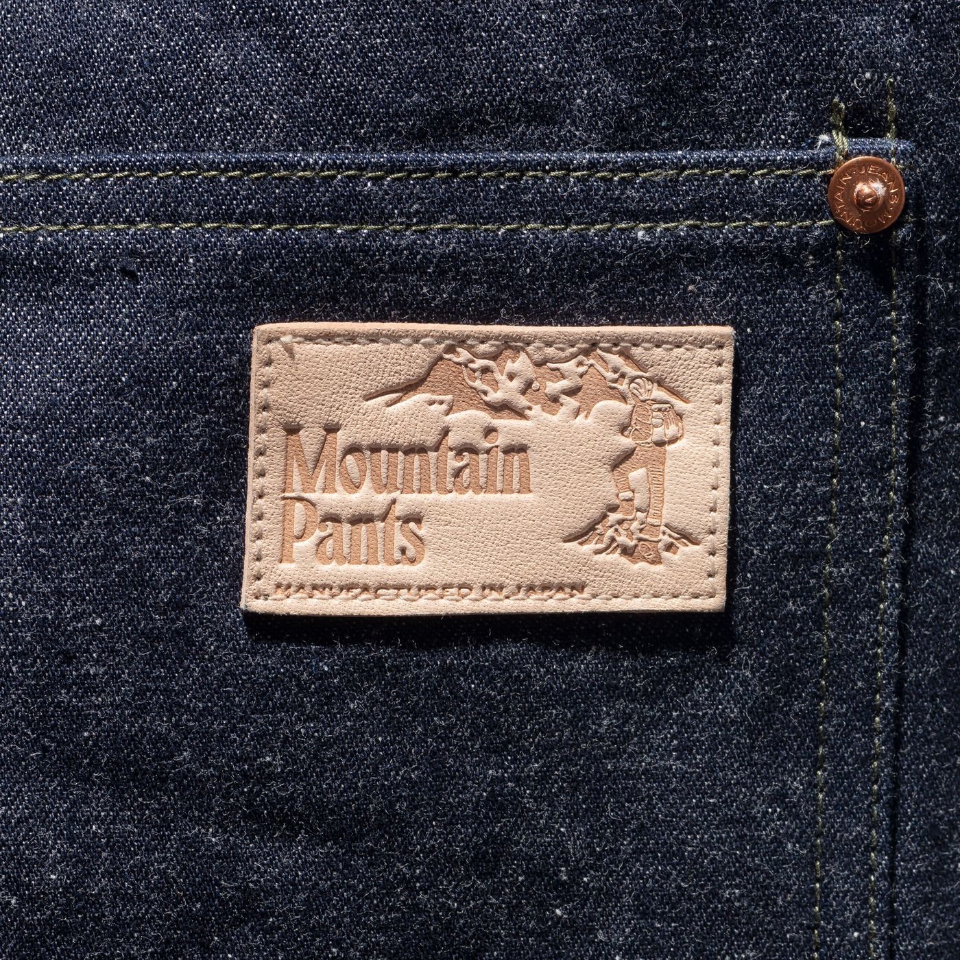 Close-up of a leather patch on dark denim, embossed with "Mountain Pants" and a mountain climber graphic, reading "Manufactured in Japan."