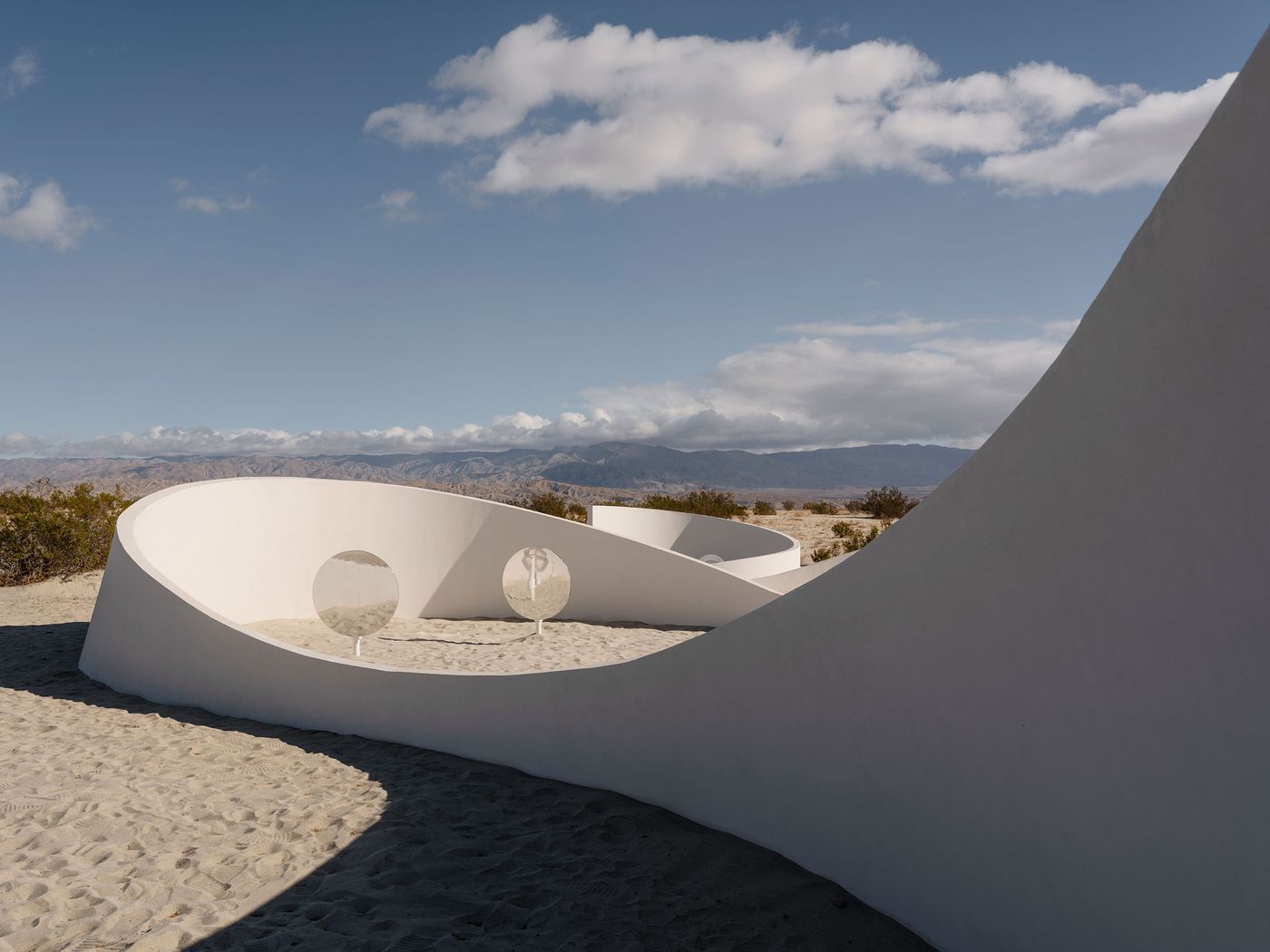 A white, curving structure with circular mirrors, set against a desert landscape and blue sky with scattered clouds.