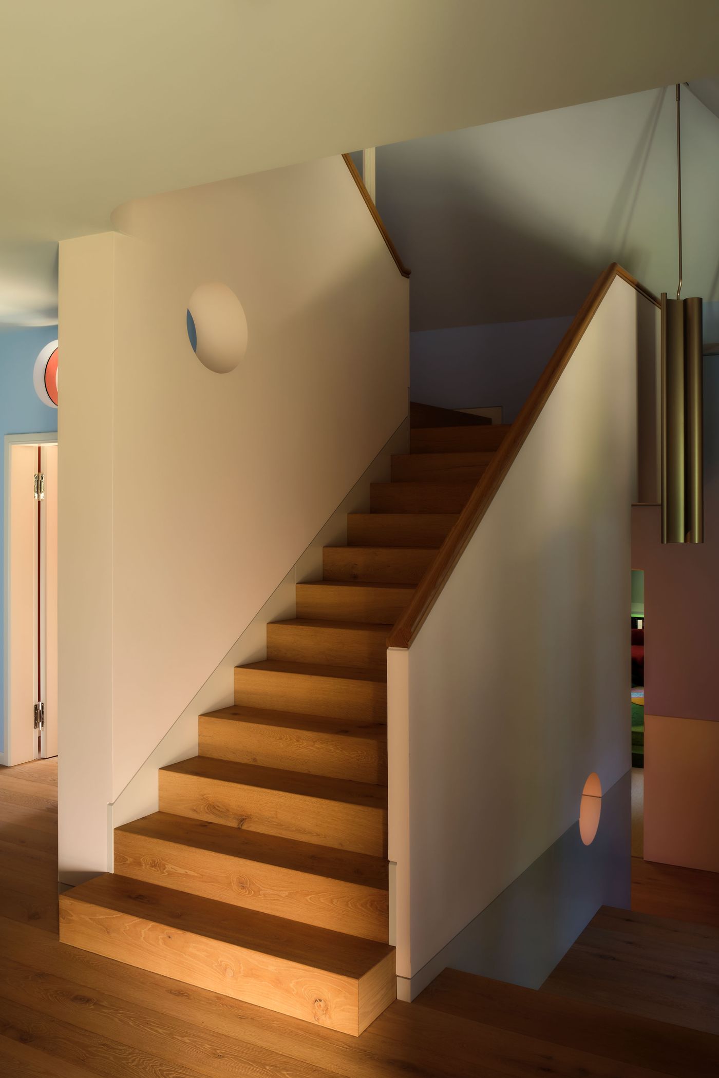 A warmly lit wooden staircase leads upward, flanked by a smooth white wall with circular cutouts and a modern pendant light overhead.