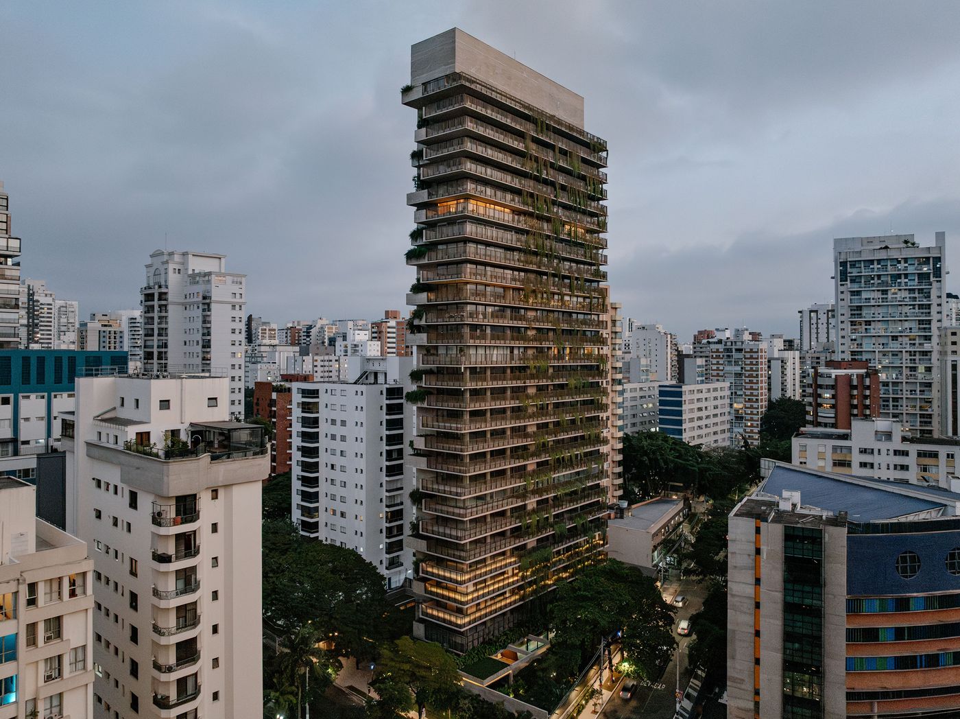 An aerial view of the Ibaté residential tower at dusk, its brutalist concrete form punctuated by glowing interior lights and extensive vertical greenery, creating a soft, inviting presence amidst the São Paulo skyline. The seamless integration of natural elements into the modern structure is a hallmark of Studio Arthur Casas's design. An aerial view of the Ibaté residential tower at dusk, its brutalist concrete form punctuated by glowing interior lights and extensive vertical greenery, creating a soft, inviting presence amidst the São Paulo skyline. The seamless integration of natural elements into the modern structure is a hallmark of Studio Arthur Casas's design.