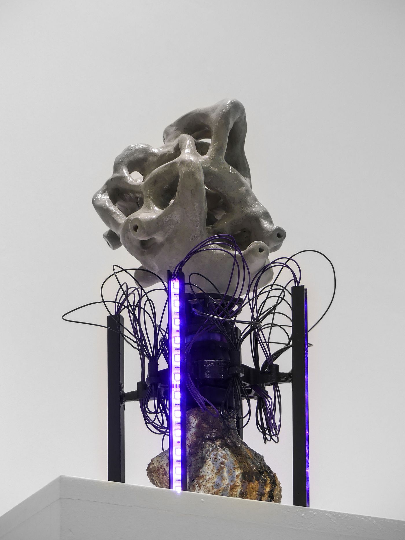 Abstract sculpture composed of a textured gray form atop a rocky base, surrounded by abstract wiring and illuminated by purple lights.