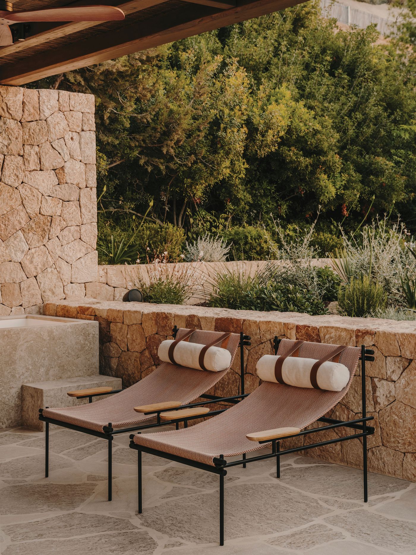 Two stylish loungers with wooden armrests and cushions sit on a stone patio, surrounded by lush greenery and warm stone walls. Two stylish loungers with wooden armrests and cushions sit on a stone patio, surrounded by lush greenery and warm stone walls.