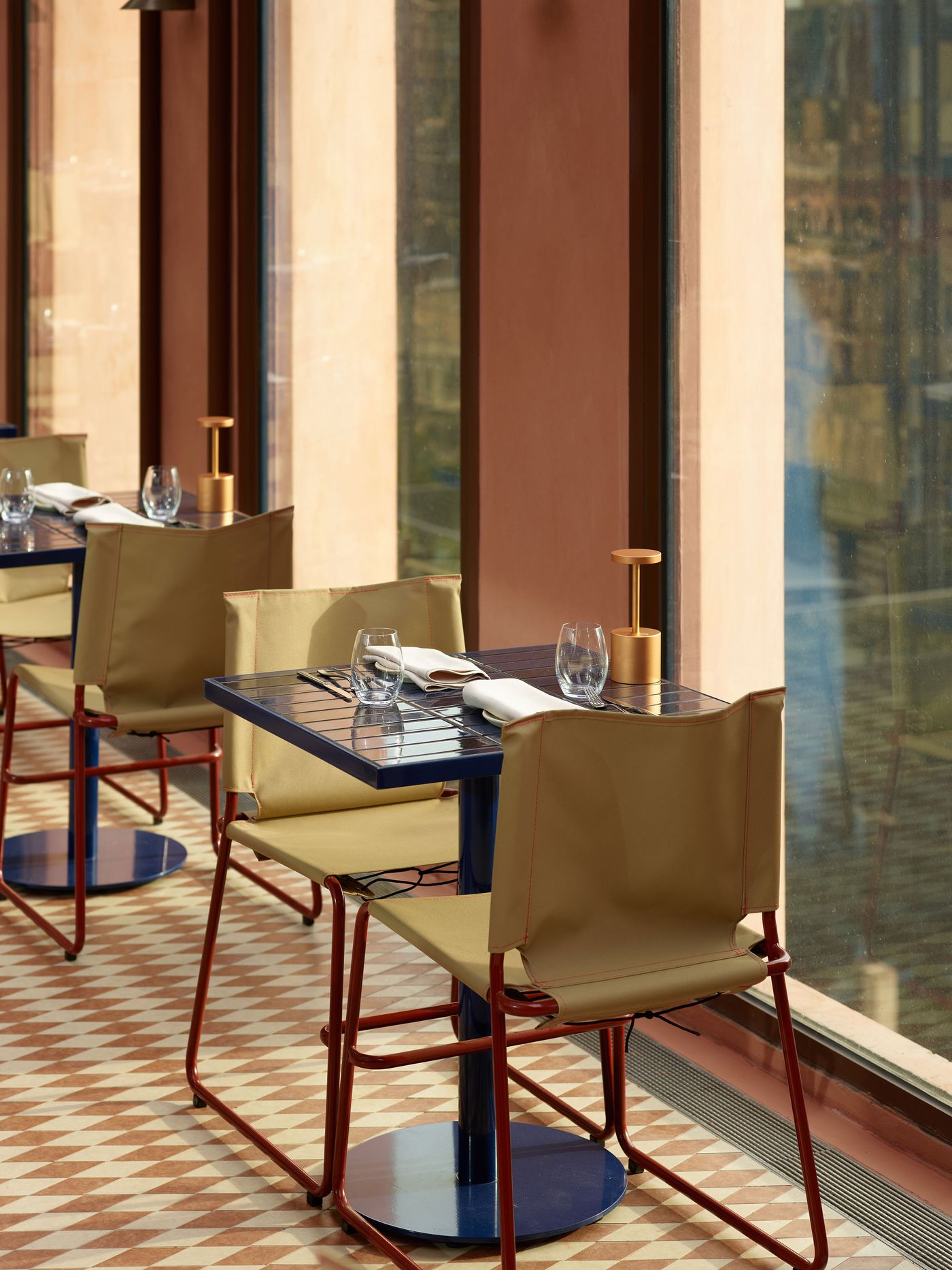 A cozy restaurant setup featuring a blue table, beige chairs, and elegant glassware, with a sunlit view of the outside. A cozy restaurant setup featuring a blue table, beige chairs, and elegant glassware, with a sunlit view of the outside.