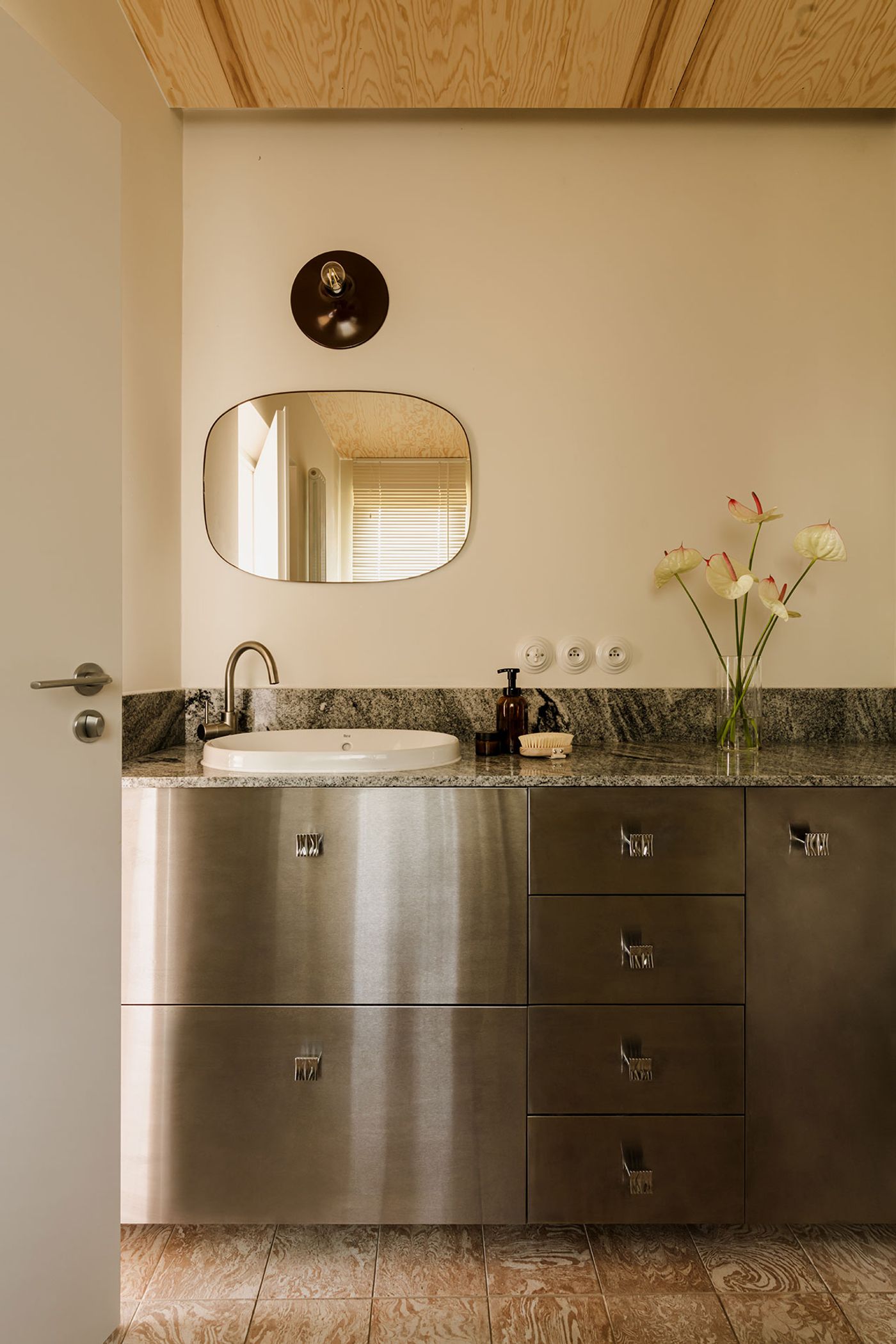 Modern bathroom featuring a sleek granite countertop, circular mirror, metallic cabinetry, and a vase of flowers for decor. Modern bathroom featuring a sleek granite countertop, circular mirror, metallic cabinetry, and a vase of flowers for decor.