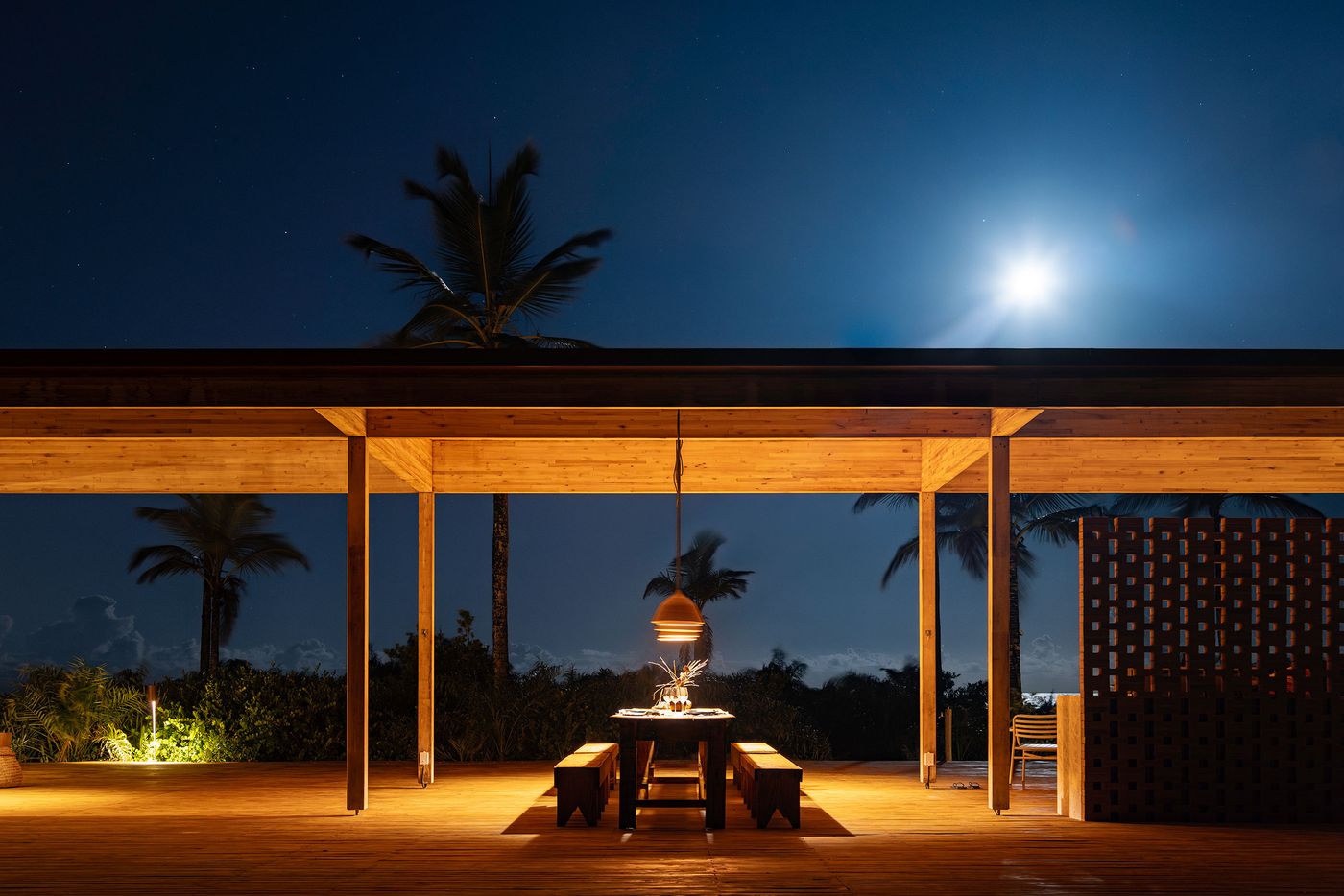 Under a bright moonlit sky, an outdoor dining setup with a rustic table and benches glows with warm light on a wooden deck. The simple, elegant structure and the silhouetted palm trees create a serene and intimate atmosphere, celebrating the tropical night. Under a bright moonlit sky, an outdoor dining setup with a rustic table and benches glows with warm light on a wooden deck. The simple, elegant structure and the silhouetted palm trees create a serene and intimate atmosphere, celebrating the tropical night.