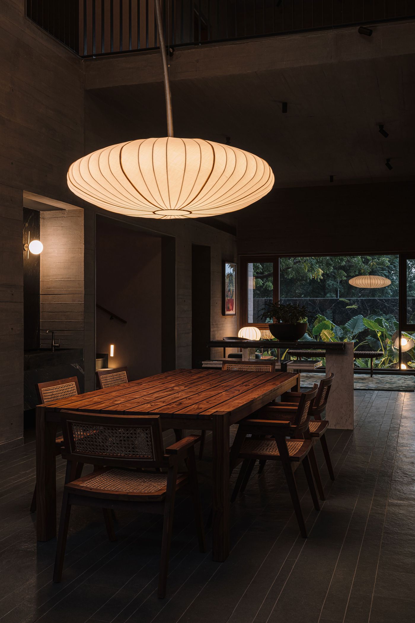 A dimly lit, atmospheric dining area, illuminated by a large, horizontal pleated pendant light that casts a warm glow over a robust wooden dining table and rattan-back chairs. The space features dark concrete walls and floor, while large windows frame a lush outdoor view, bringing a touch of nature into the sophisticated, modern interior.