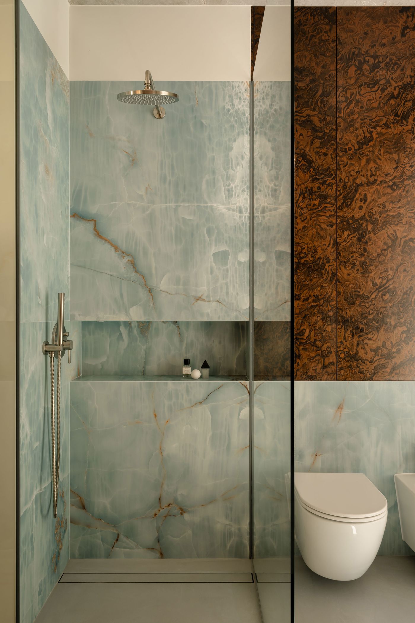 The walk-in shower showcases an elegant backdrop of veined blue onyx and walnut cabinetry, with sculptural matte fixtures adding a refined, contemporary edge to the richly textured surfaces. The walk-in shower showcases an elegant backdrop of veined blue onyx and walnut cabinetry, with sculptural matte fixtures adding a refined, contemporary edge to the richly textured surfaces.