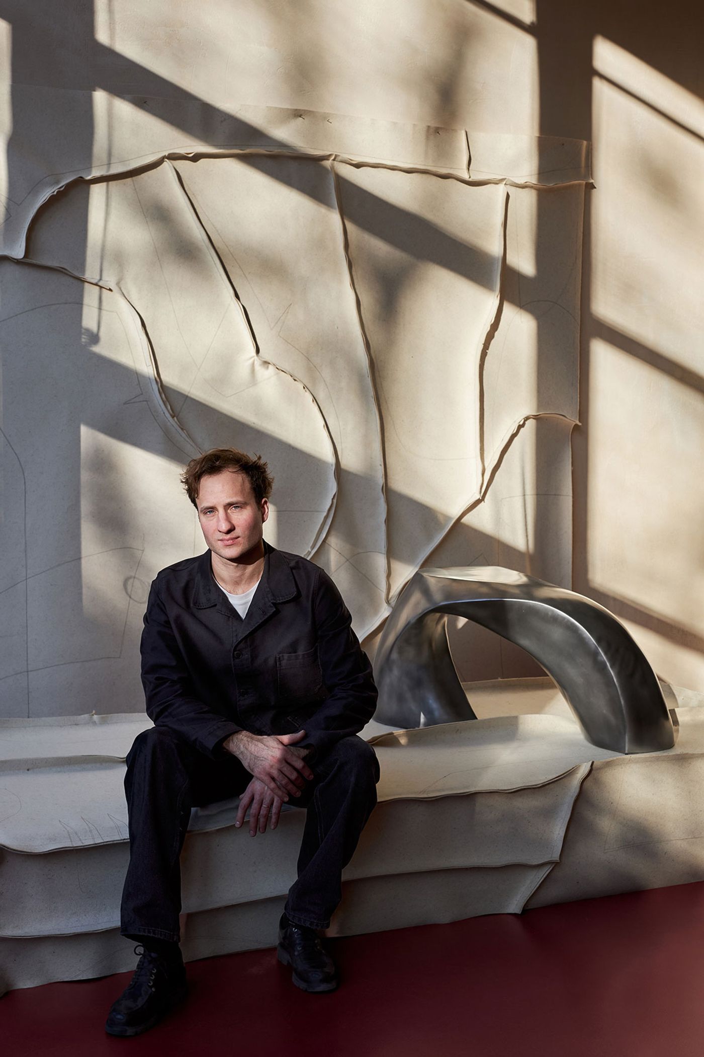 A person in dark clothing sits on a sculptural bench against a textured wall, illuminated by geometric shadows from large windows.