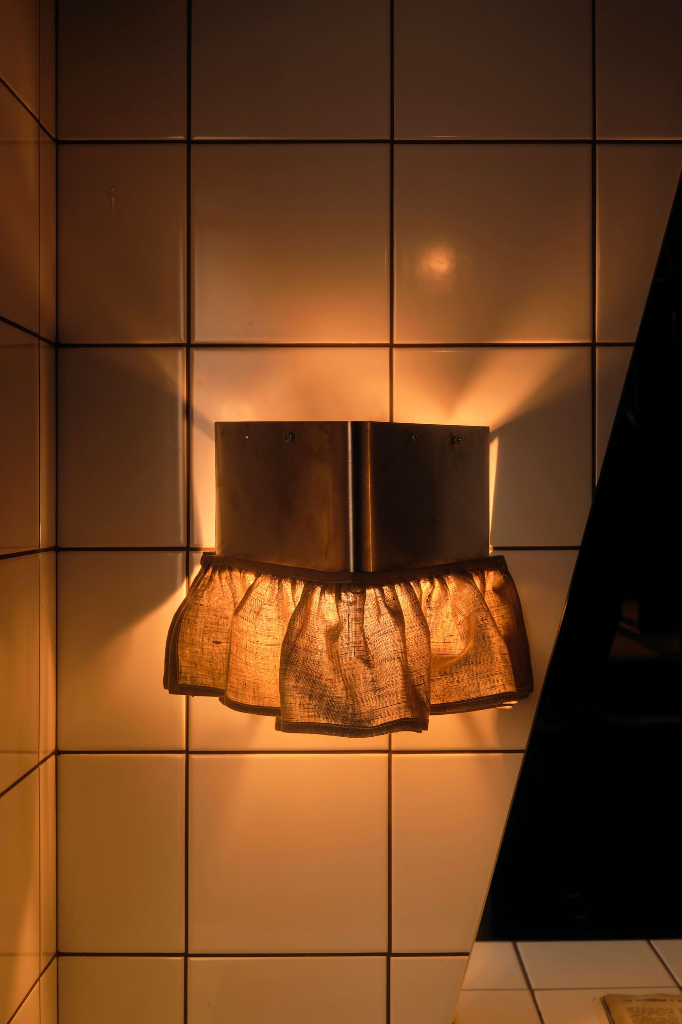A distinctive wall sconce with a metallic, box-like top and a ruffled fabric skirt illuminates a tiled wall at The Roses of Elagabalus. The warm light creates dramatic shadows and highlights the texture of the fabric, contributing to the eclectic and luxurious vintage-retro aesthetic of the space.