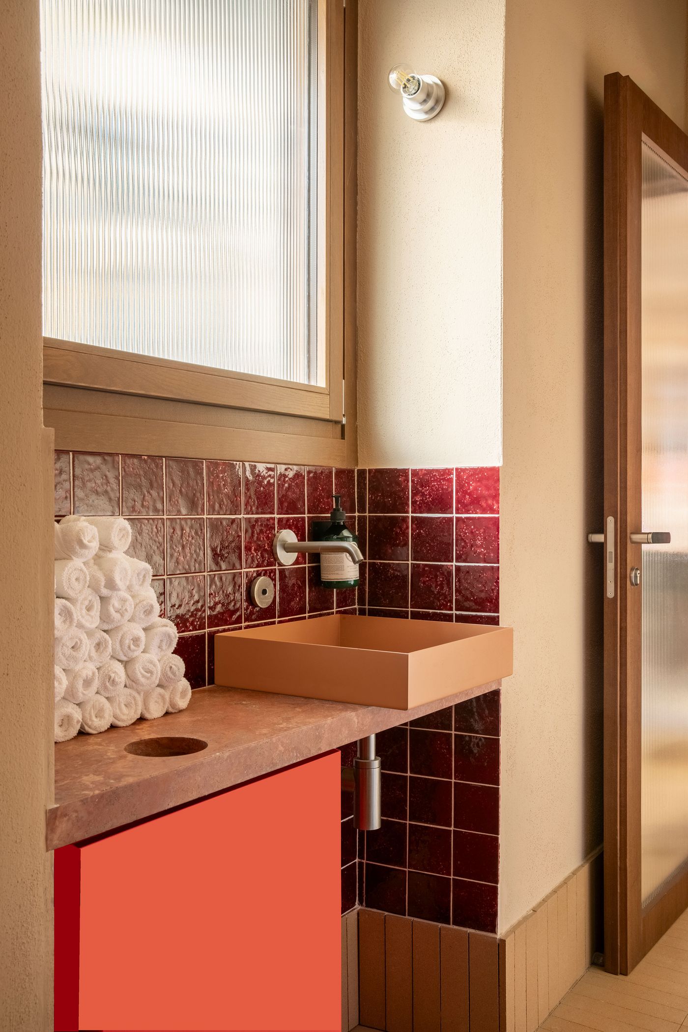 A vibrant and modern restroom interior at Santo Palato by Naessi Studio, featuring a terracotta tiled backsplash and a minimalist, light-colored sink basin. Rolled white towels are neatly stacked, and a window with fluted glass adds a touch of privacy. The bold color scheme and clean lines create a contemporary yet warm space.