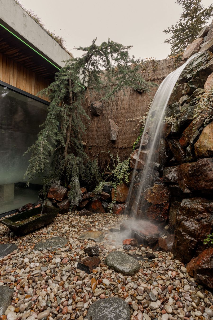 A tranquil indoor waterfall cascades over rocks into a pebbled basin, surrounded by greenery and modern architecture. A tranquil indoor waterfall cascades over rocks into a pebbled basin, surrounded by greenery and modern architecture.